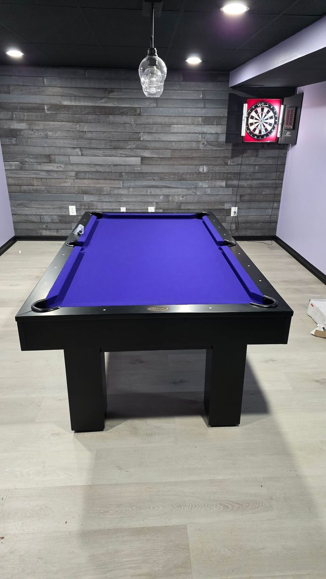 A pool table with bright purple felt and a black frame in a room with gray wood-paneled walls and a dartboard.