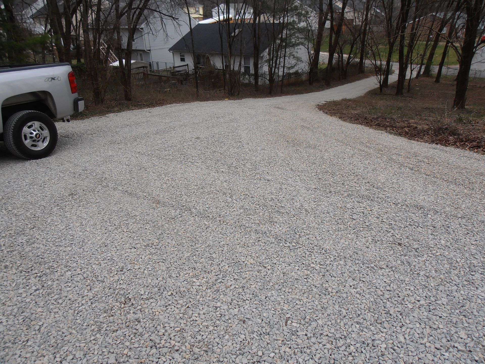 Silver pickup truck parked on gravel driveway leading to a house in a wooded area.