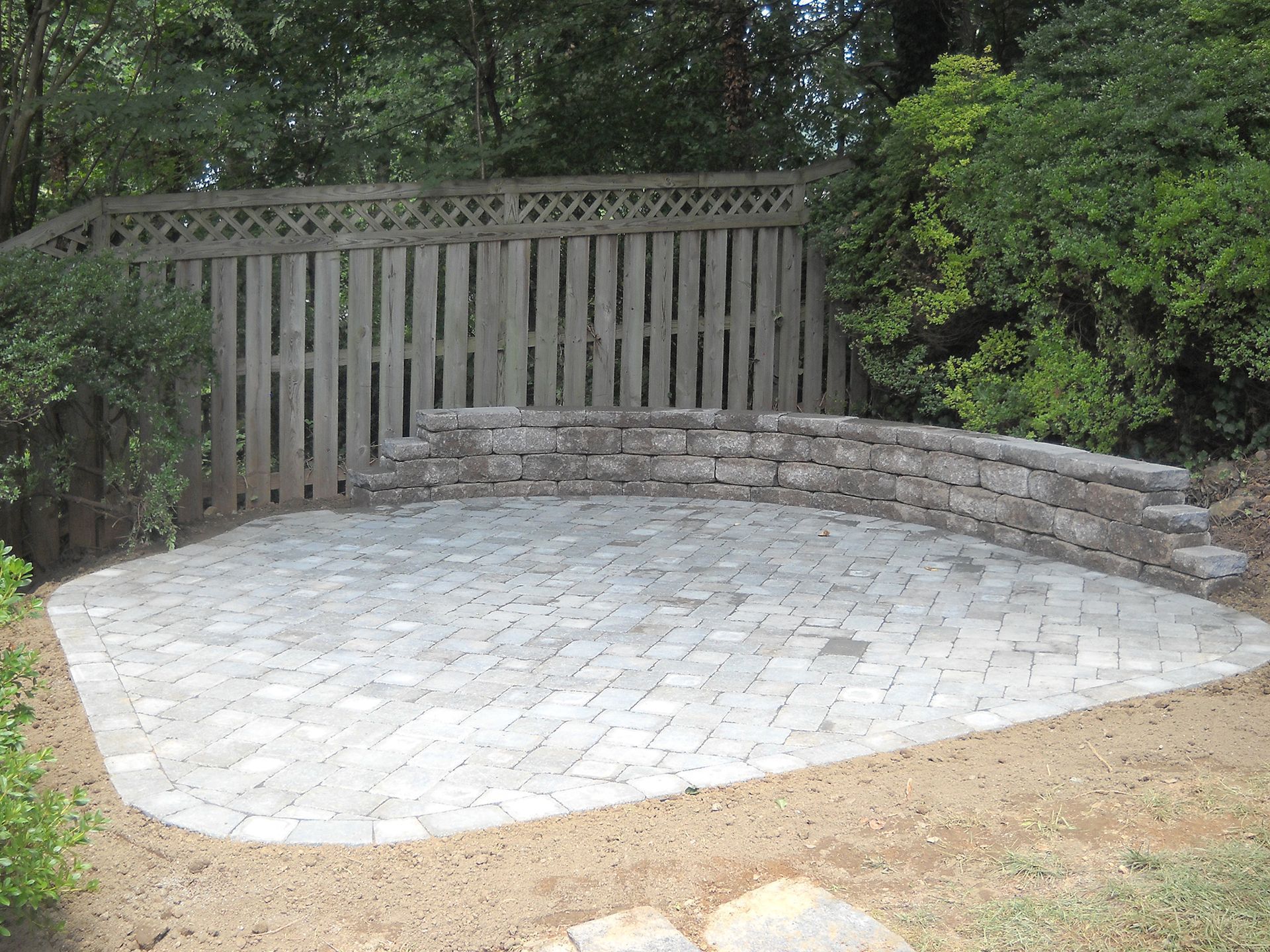 A stone patio with a curved retaining wall and a wooden fence backdrop in a yard.