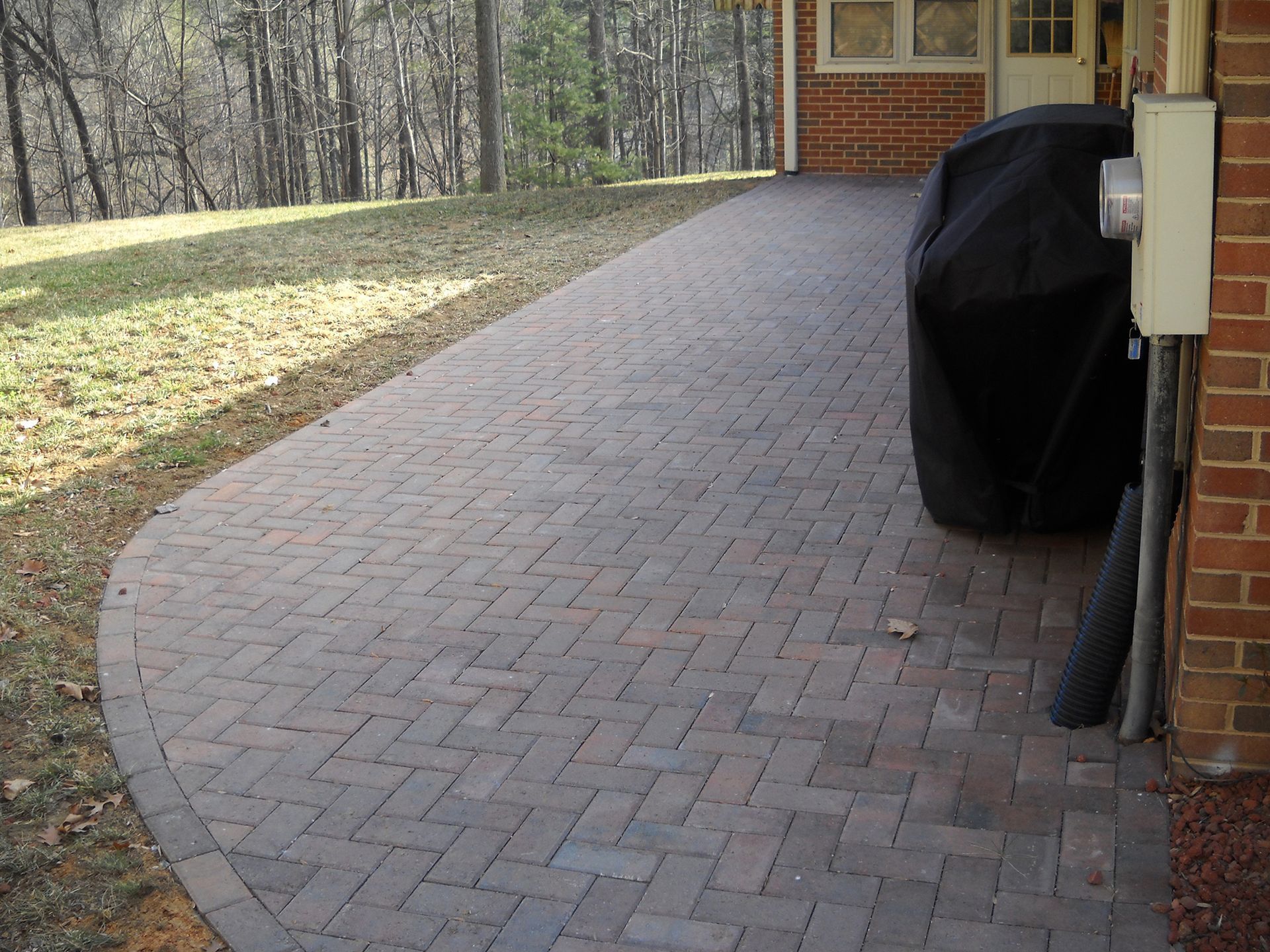 Brick patio next to a grassy lawn, leading to a house. A covered grill and electrical box are visible.
