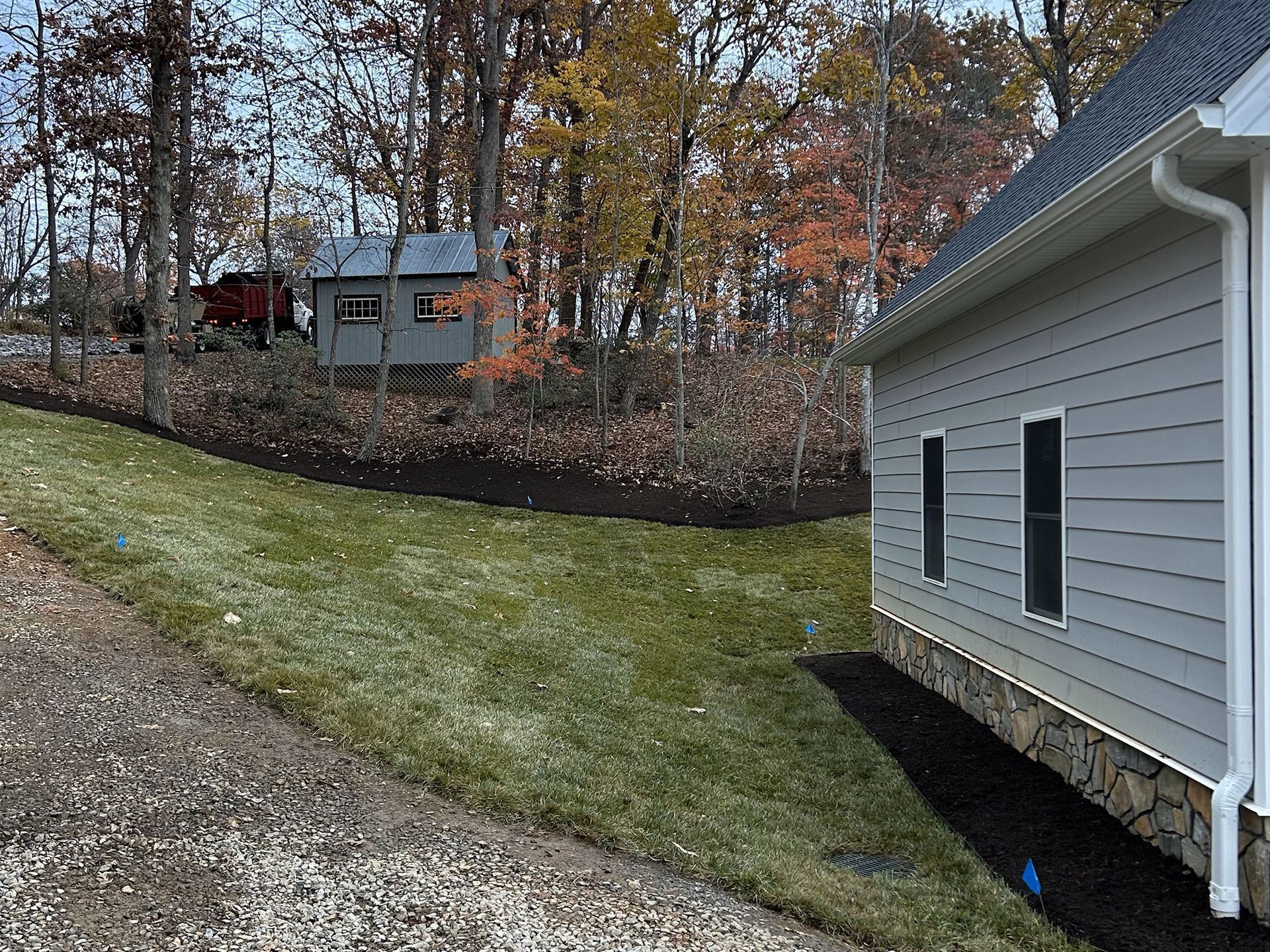 Exterior view of a house with a shed on a hill in a wooded area. Light blue siding, green grass.