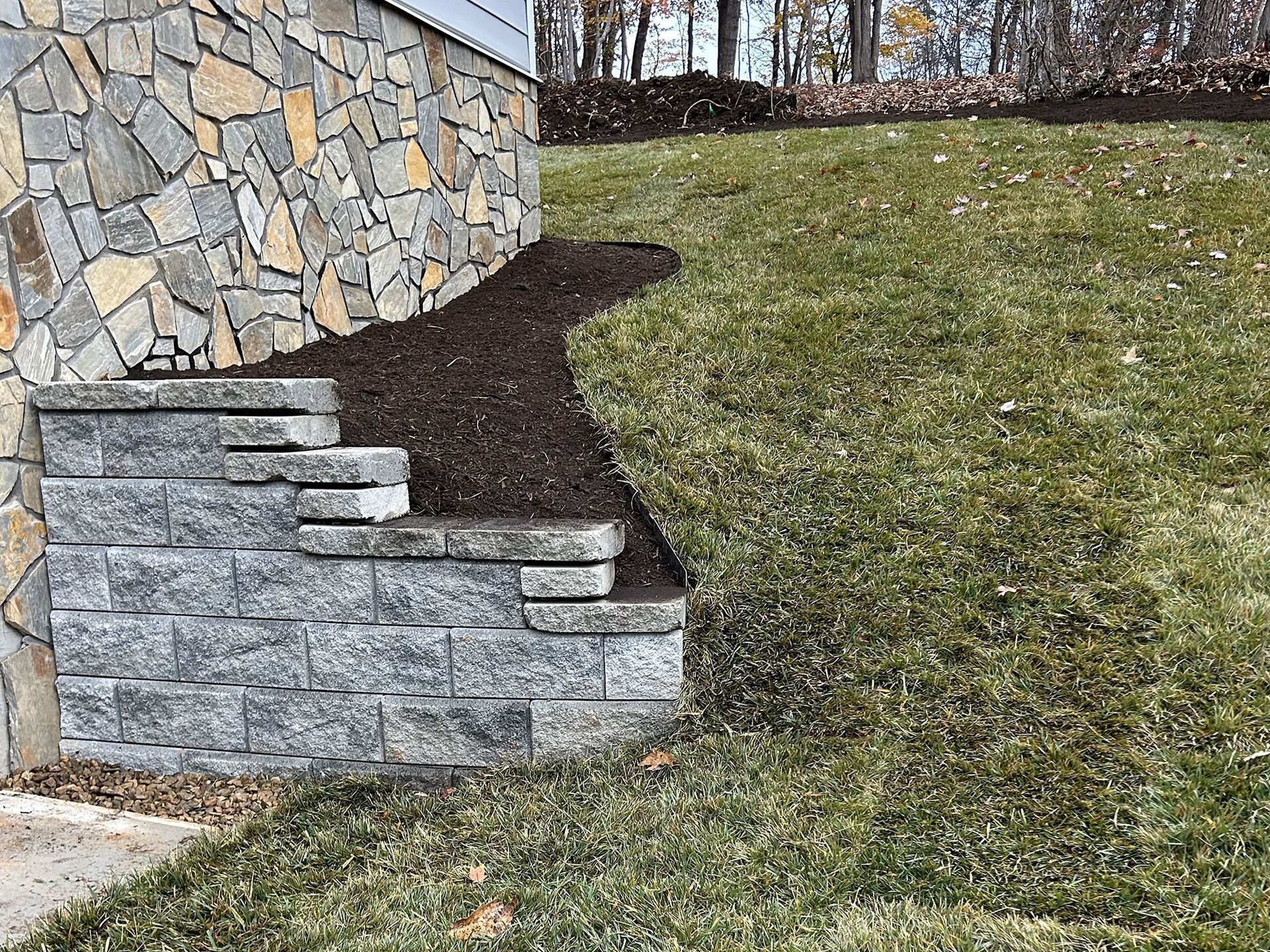 Stone retaining wall with steps next to a house with a stone facade and sloped lawn.