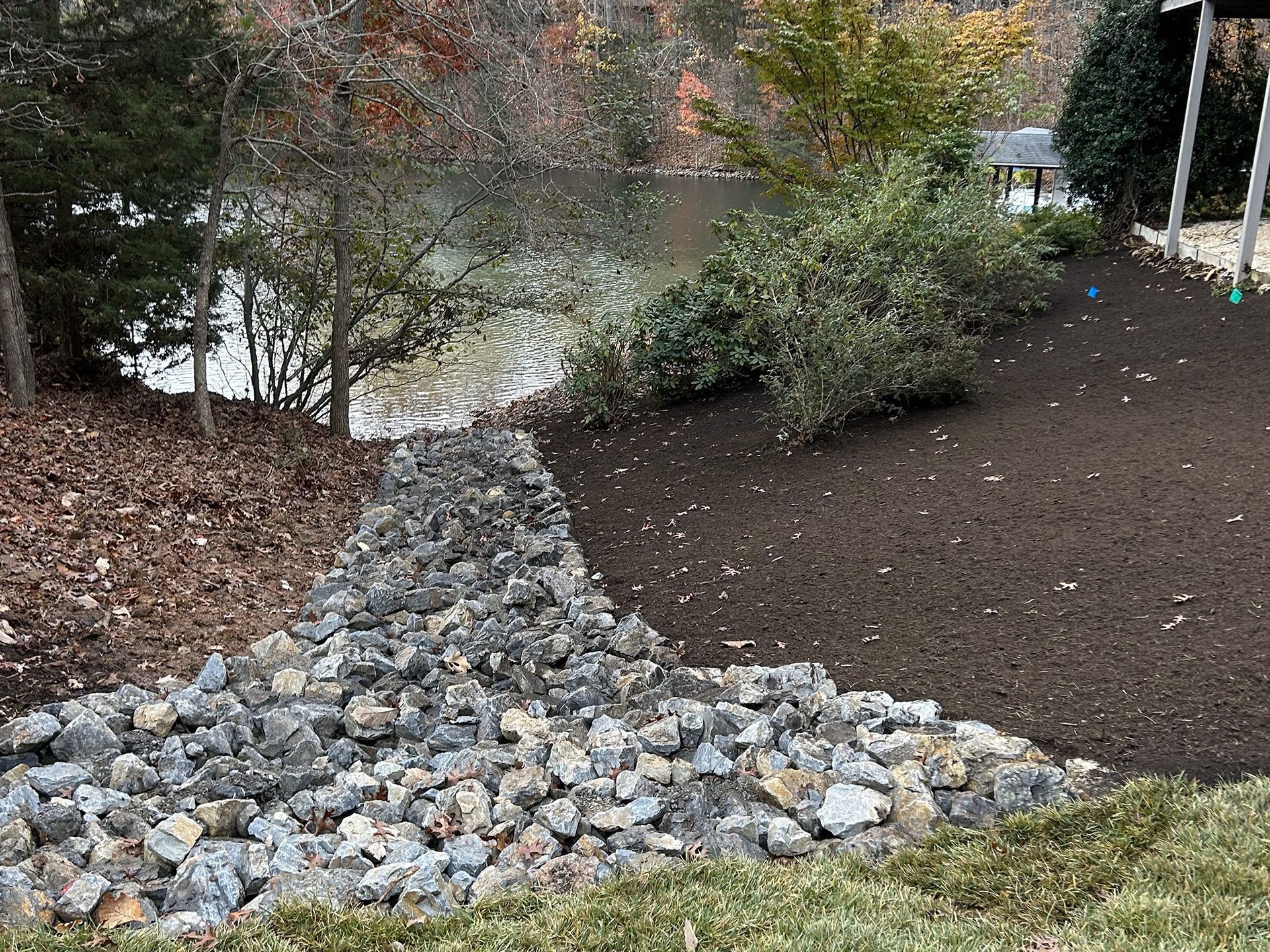 Rocks and dirt border a lake. Trees and bushes are nearby. The ground is covered with leaves.