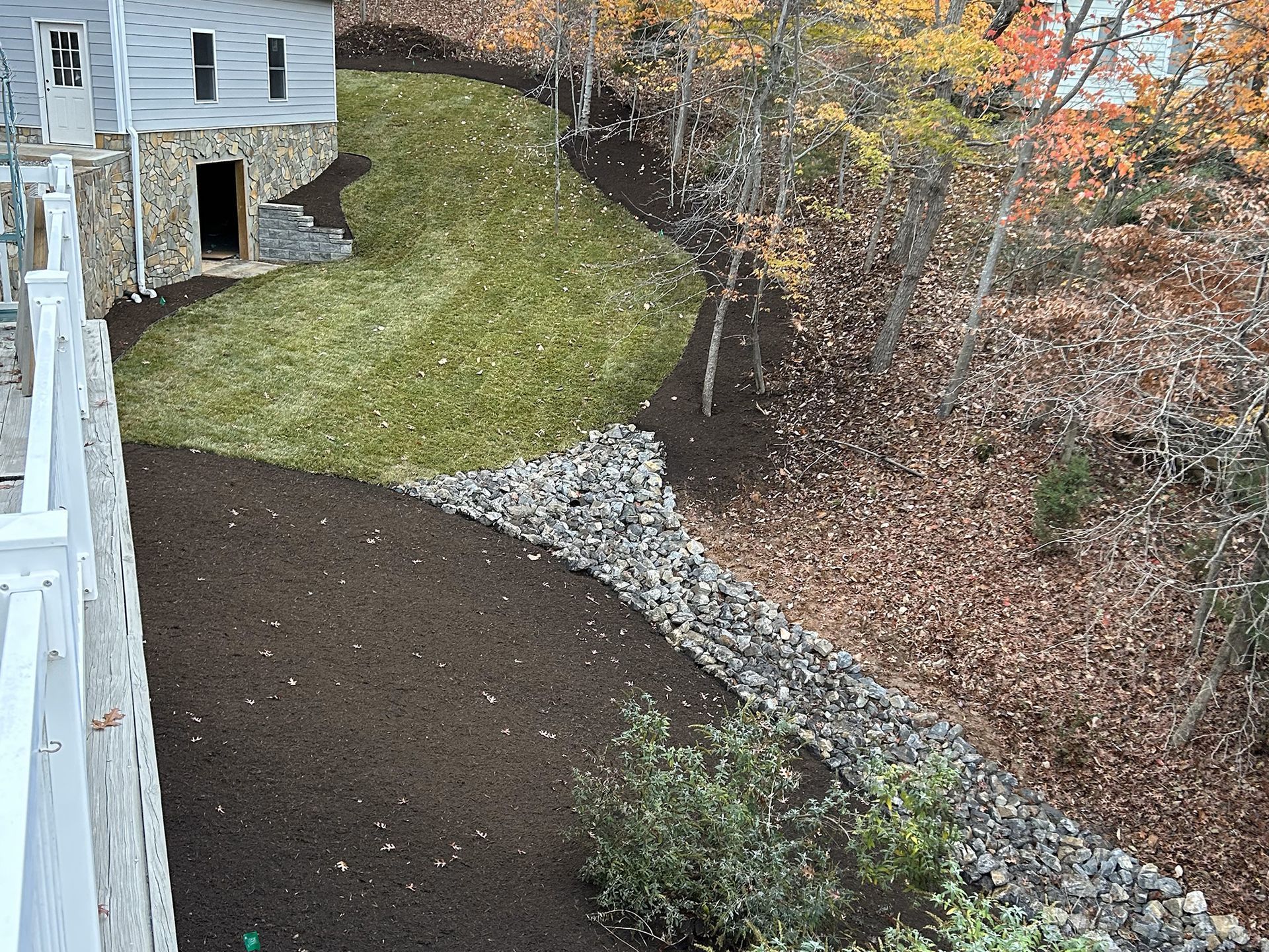 Yard with grass, mulch, and a rock-lined border. A house and trees are in the background.