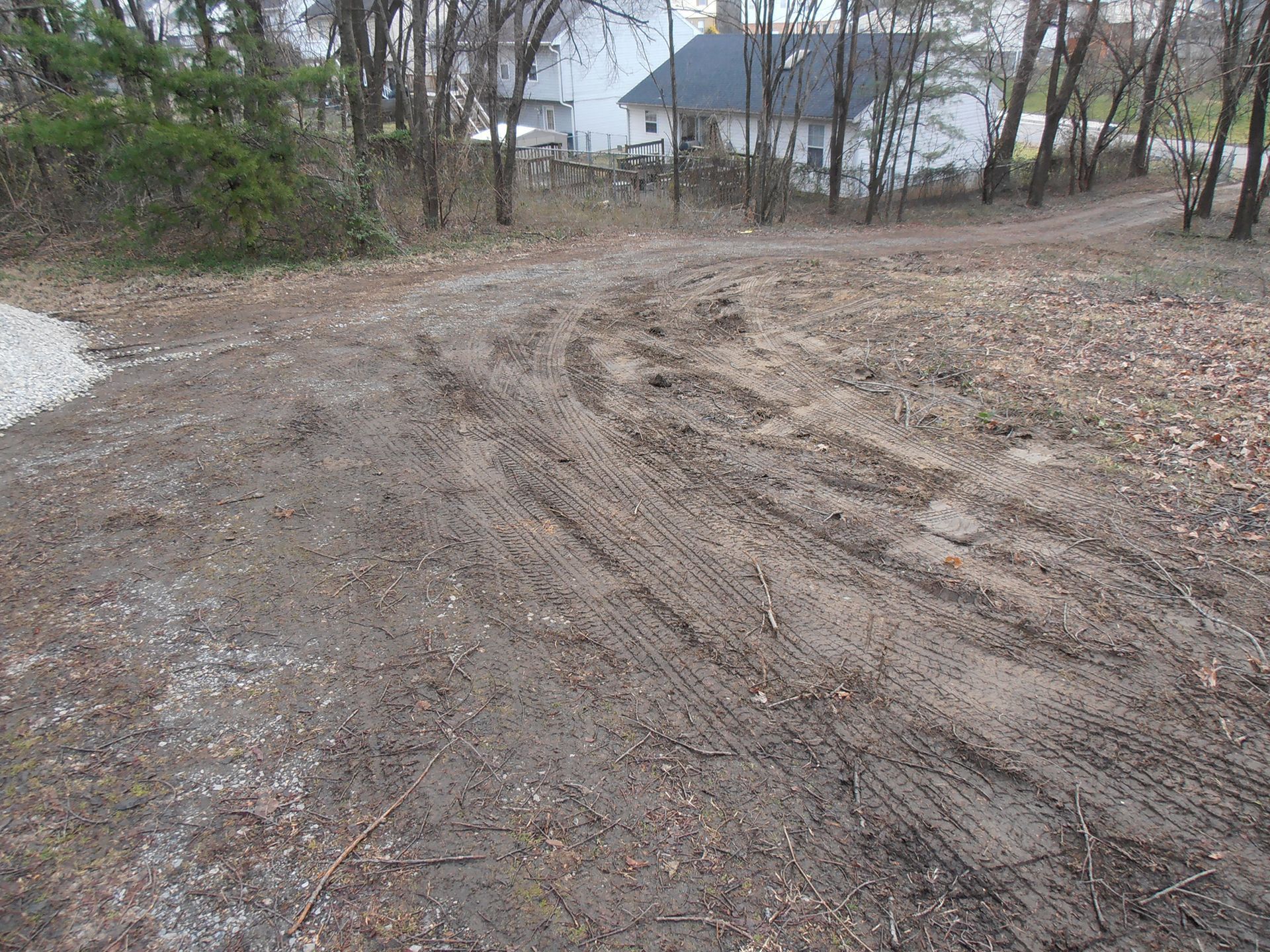 Muddy dirt road with tire tracks; trees and buildings in the background.