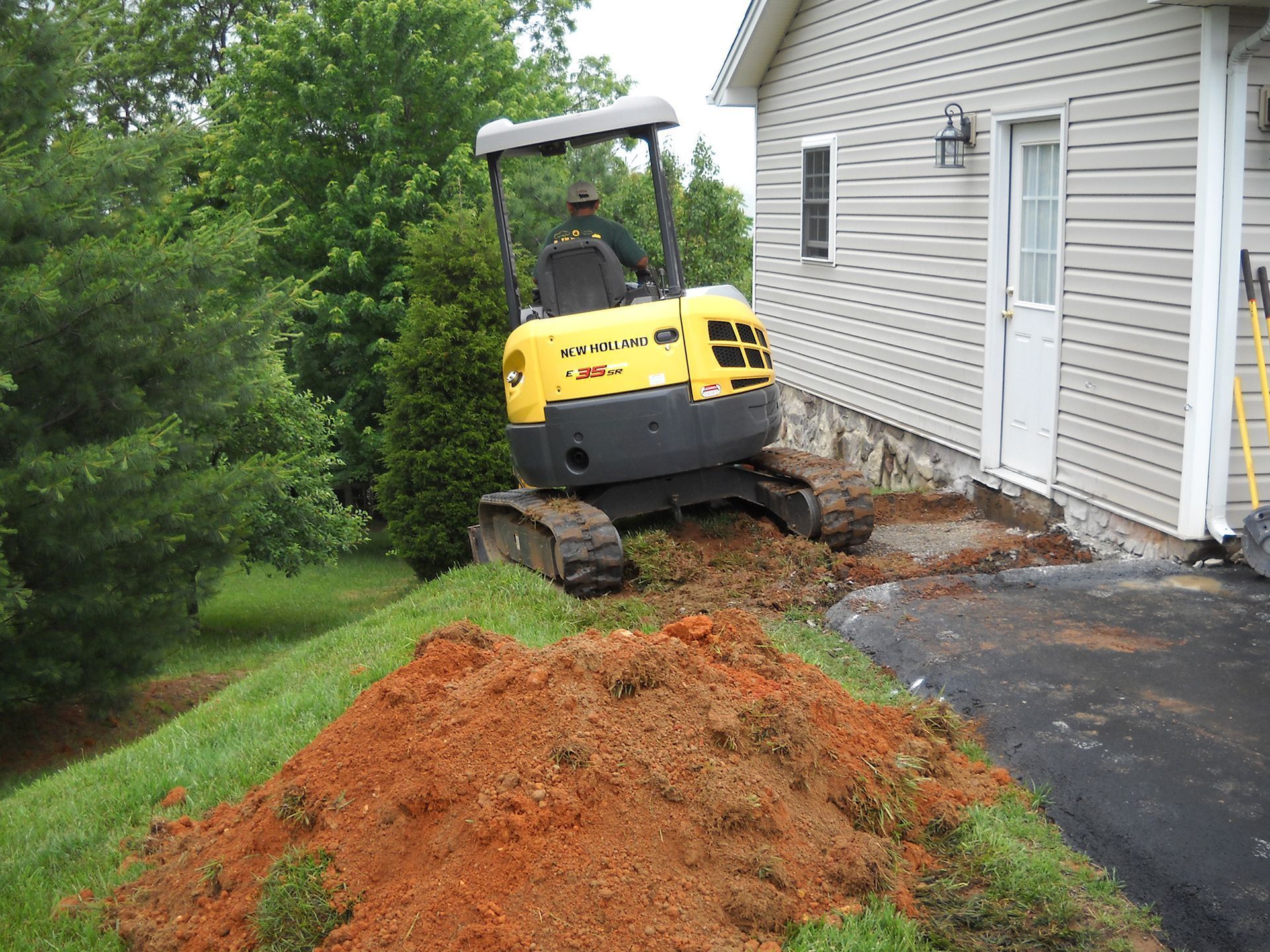 Yellow excavator digging near a house, creating a pile of red dirt on a grassy hill.