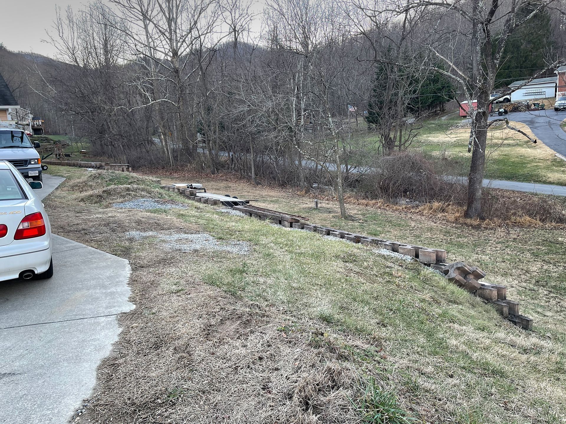 A white car parked next to a paved driveway and grassy area with a low rock wall. Trees in the background.