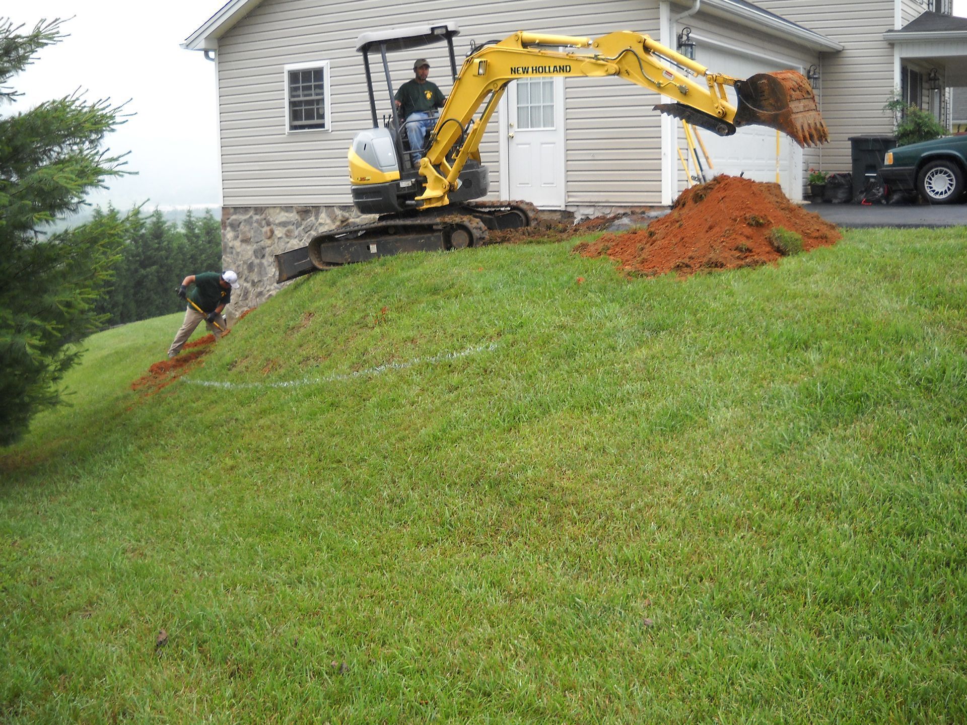 Yellow excavator digging dirt on a grassy hill; worker marking a line. House in the background.