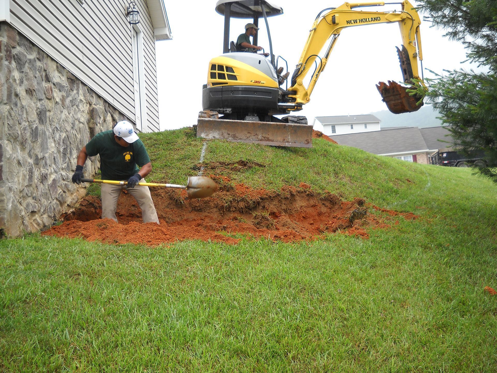 A worker shovels dirt near a house foundation while an excavator digs into a hill.