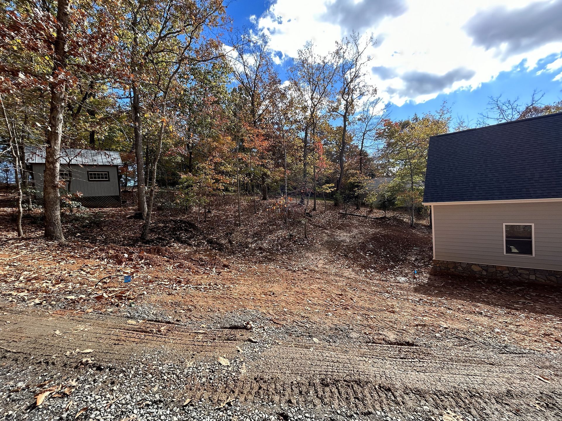 Dirt lot with bare trees, a small shed, and a partially constructed building under a partly cloudy sky.