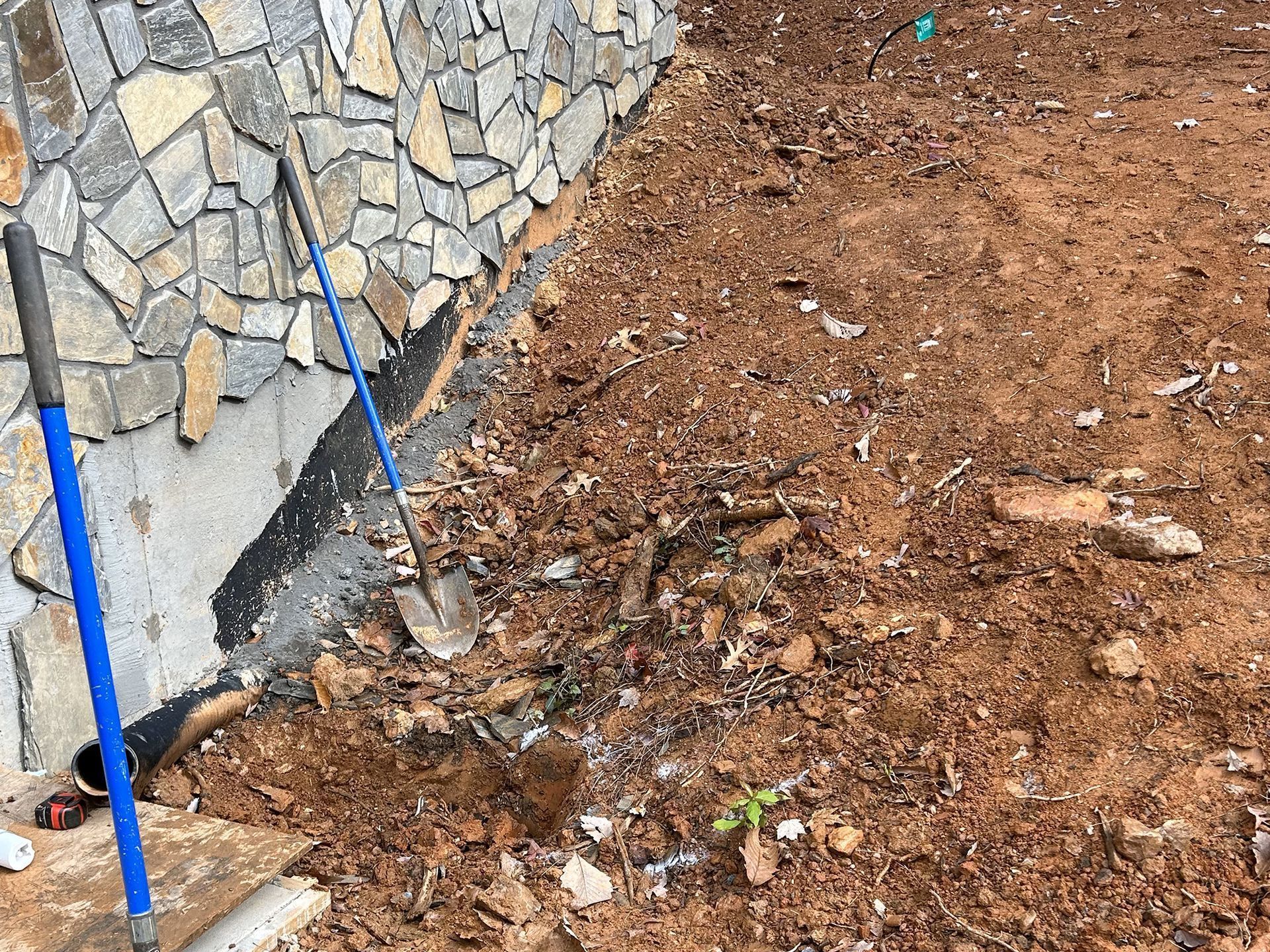 Stone wall next to dirt area with blue handled tools. Some exposed foundation is visible.