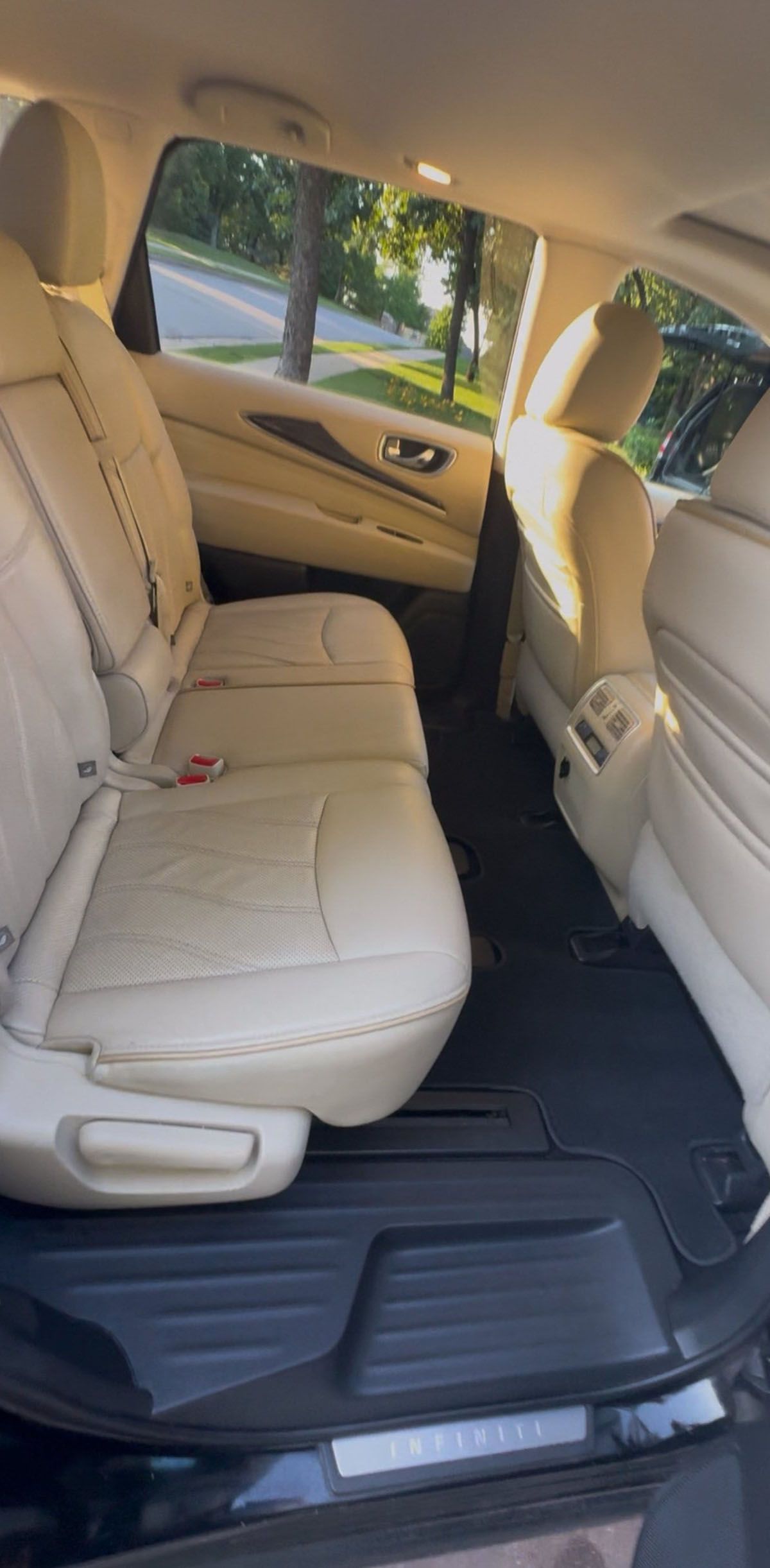 Interior view of a vehicle's back seat. Light-colored leather seats, black floor mats, and a window with a view of trees.