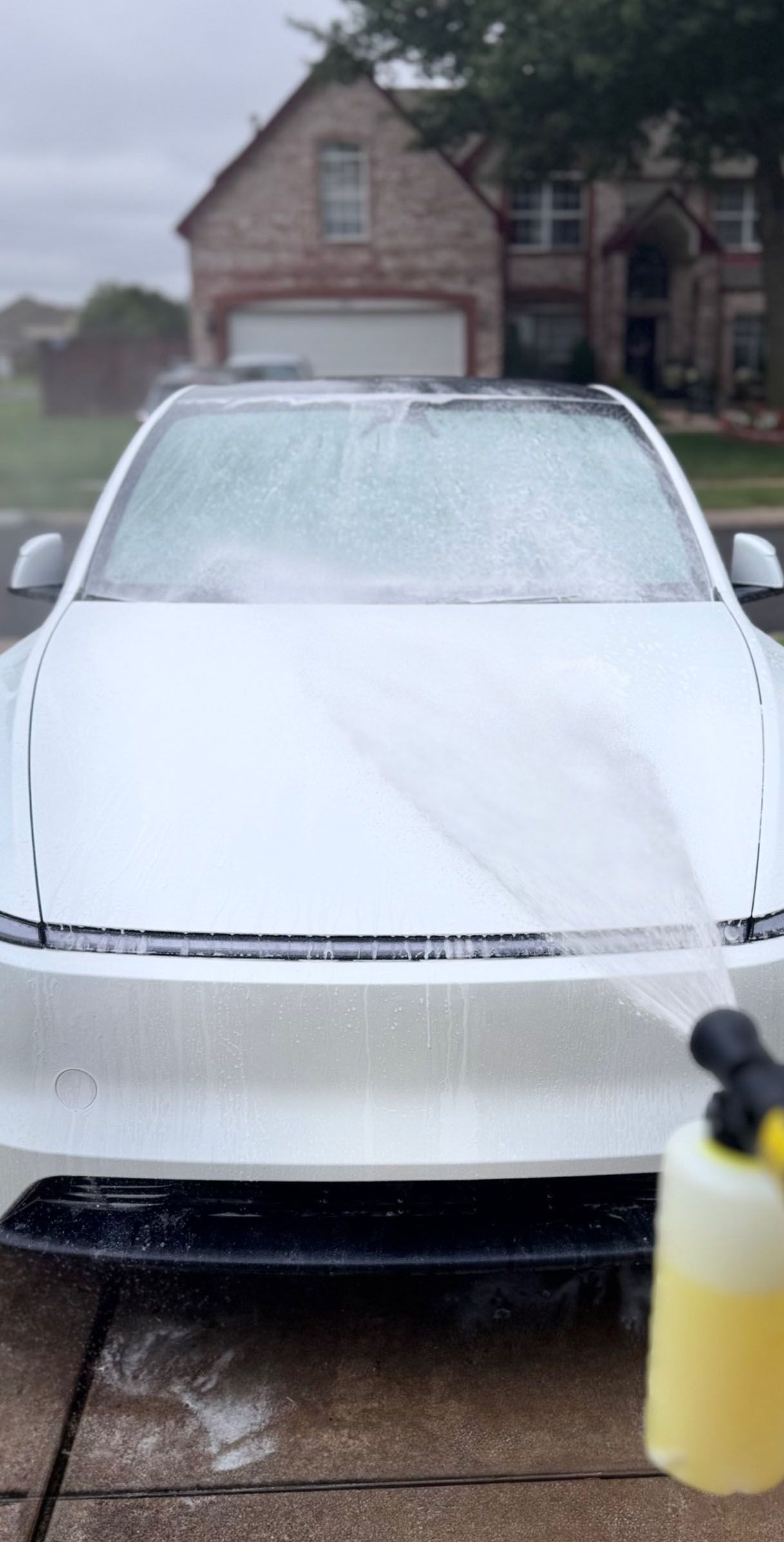 A white car is being washed with foamy soap. The house is in the background on a cloudy day.