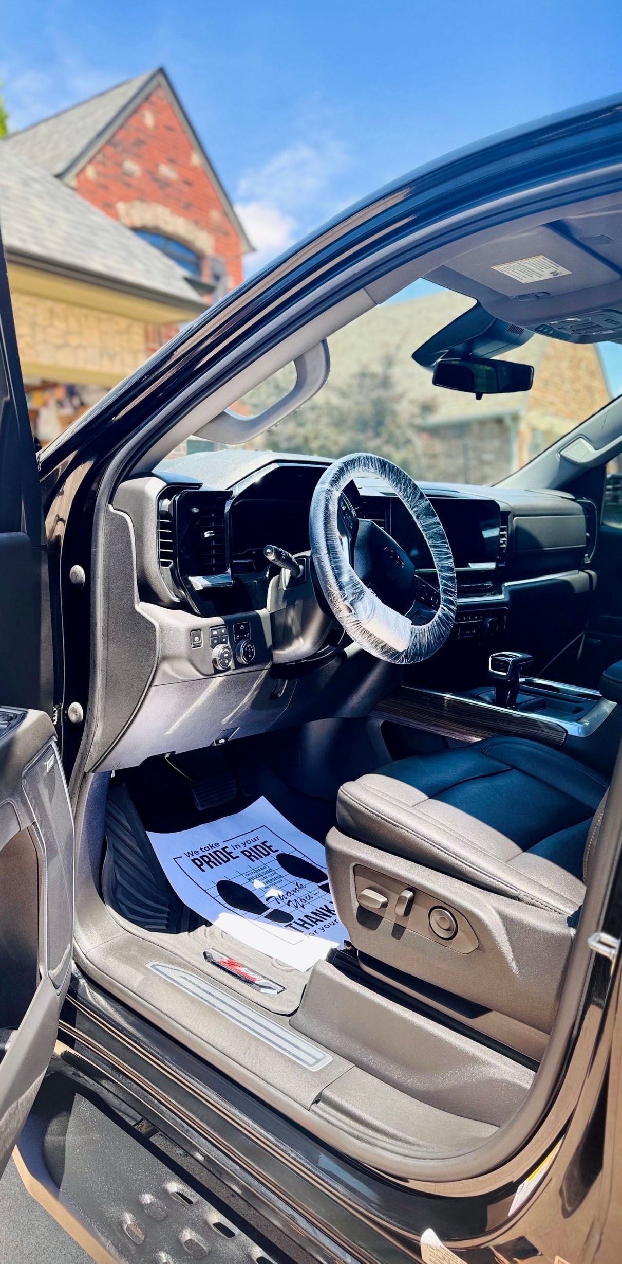 Interior of a black pickup truck with a steering wheel cover, open door, and a building in the background.