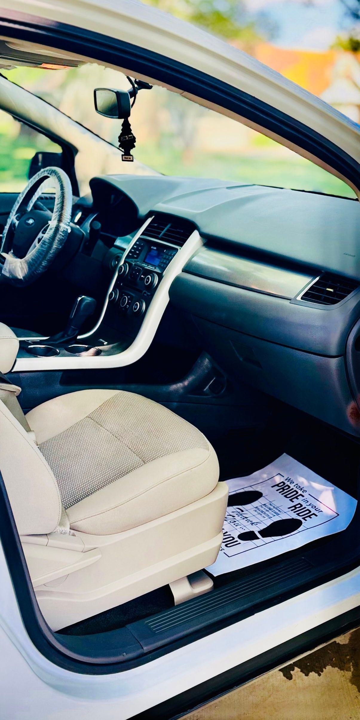 Interior view of a white car, showing the dashboard, steering wheel, seats, and a paper mat on the floor.