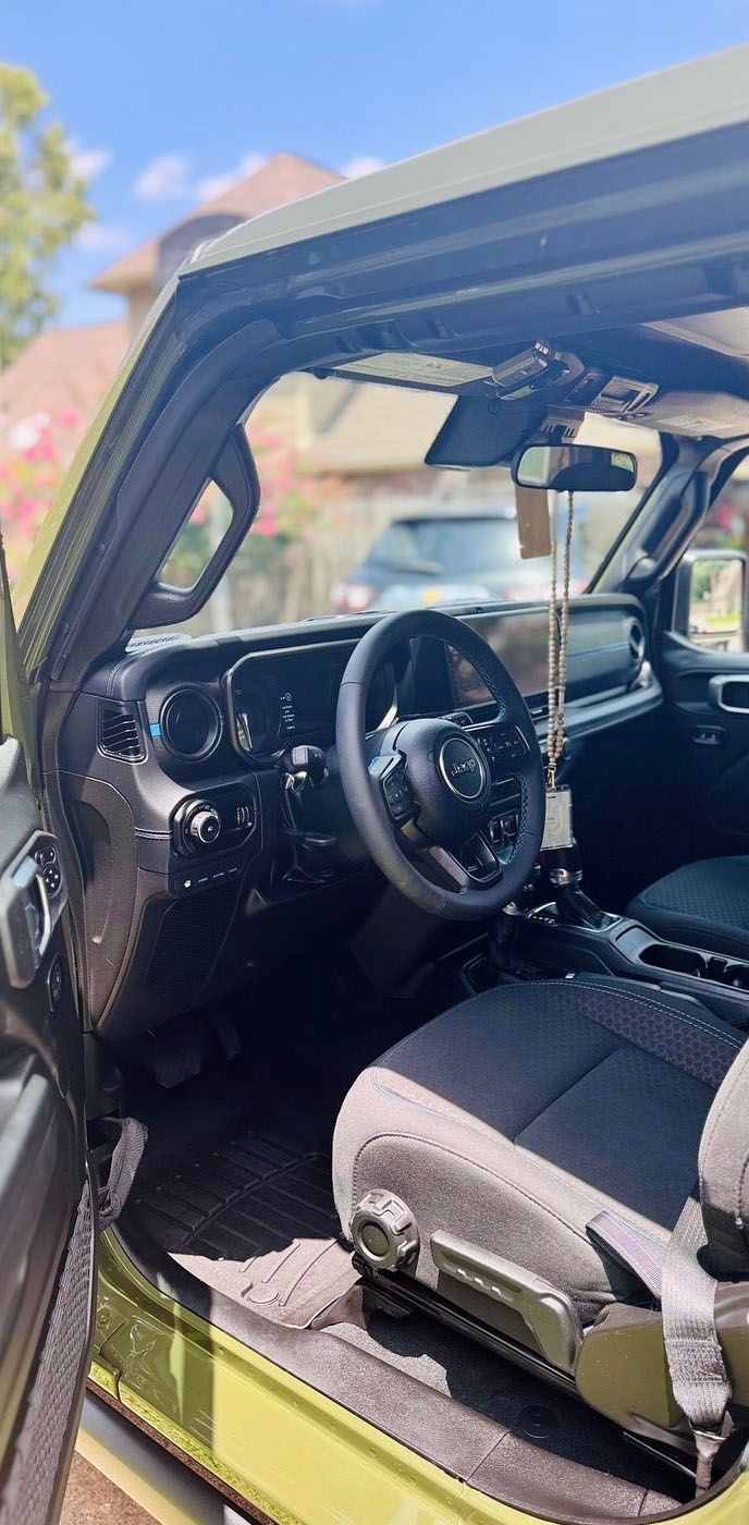 Interior view of a green Jeep Wrangler. Steering wheel and dashboard are visible. A black seat fills the foreground.