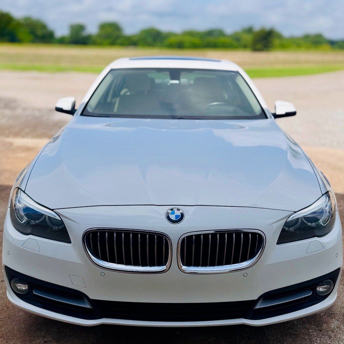 White BMW sedan parked outdoors, facing the viewer. The sky and a field are in the background.