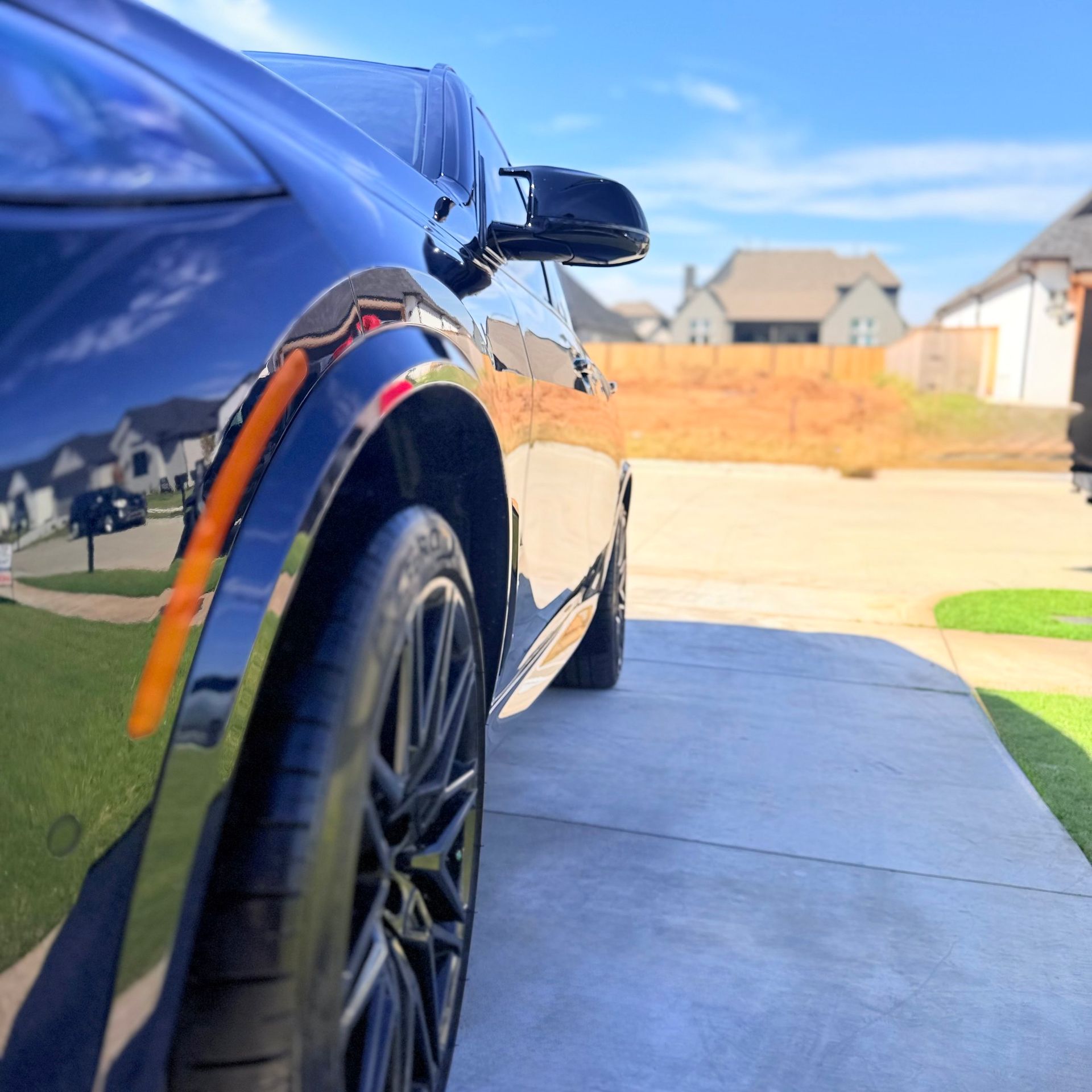 Black car parked on a concrete driveway, reflecting the sunny day, neighborhood in background.