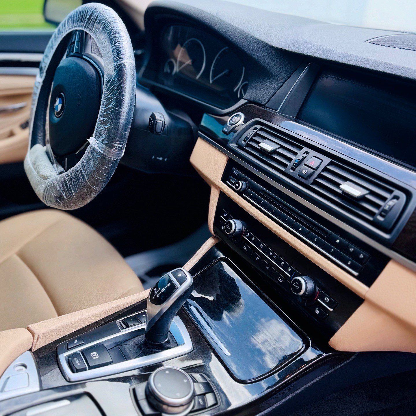 Interior of a tan and black BMW car, dashboard, steering wheel covered in plastic.