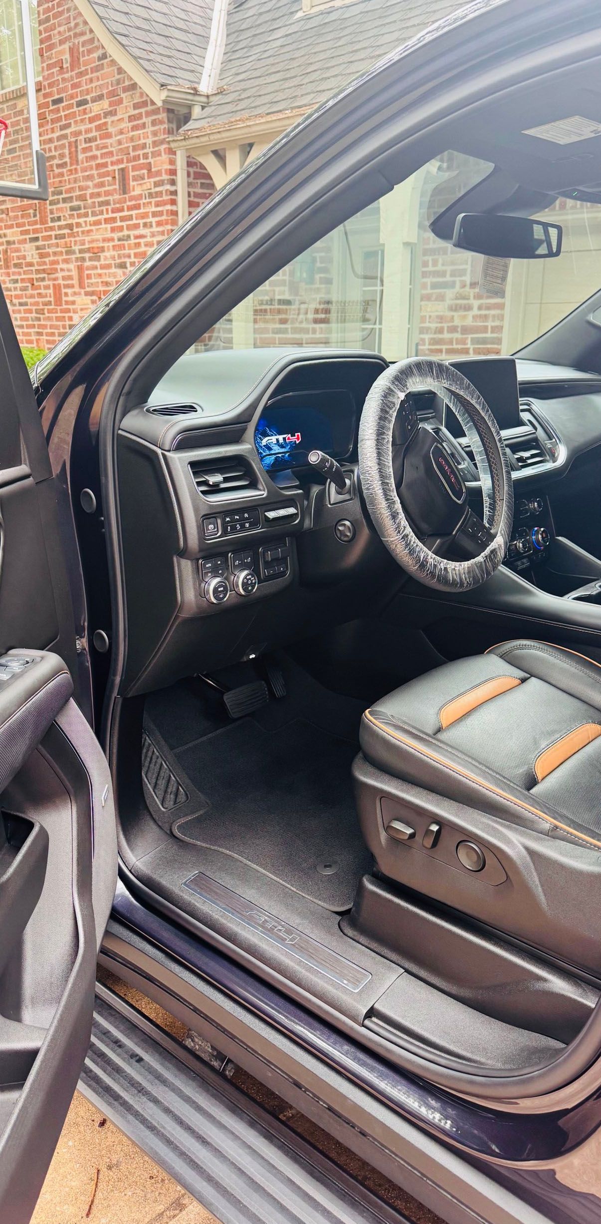 Interior view of a dark SUV. Black leather seats with tan accents, steering wheel cover. Open door reveals brick house.