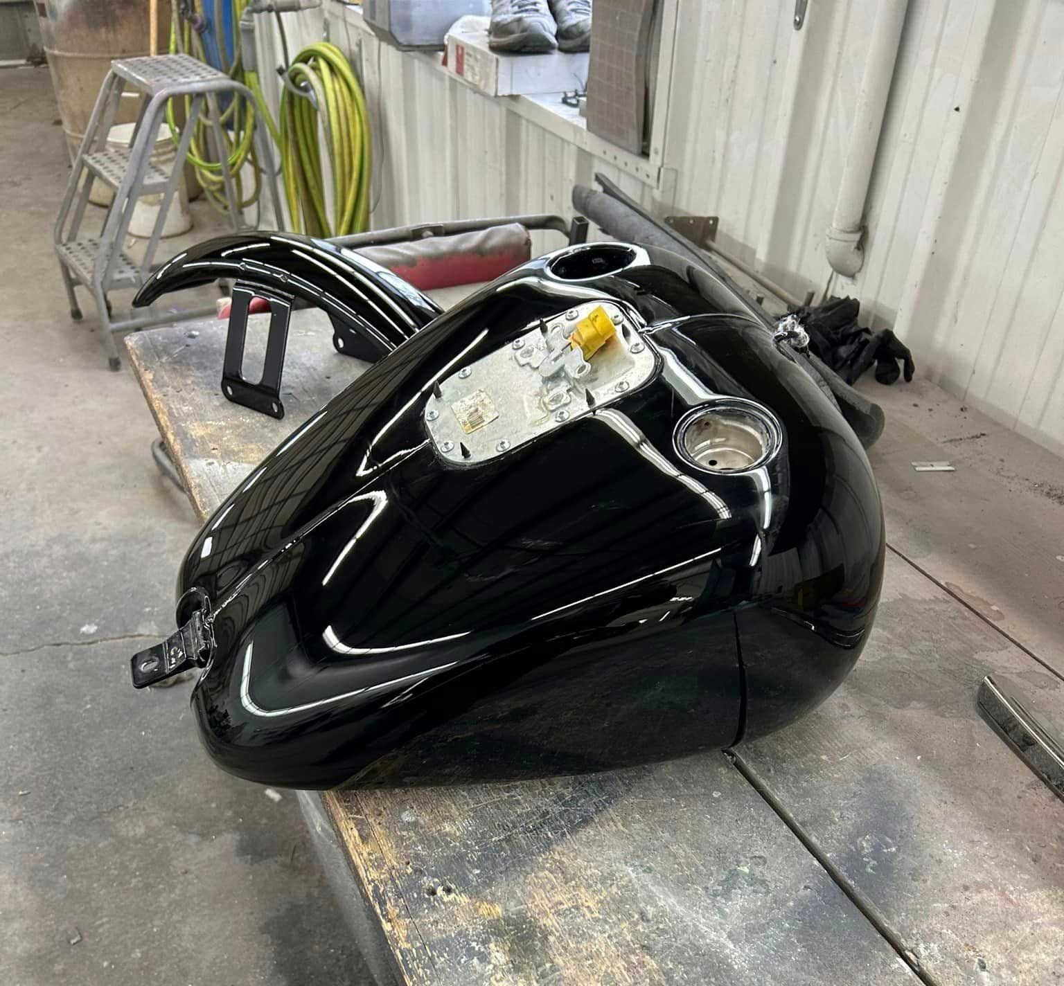 Black motorcycle gas tank with glossy finish, resting on a workbench.