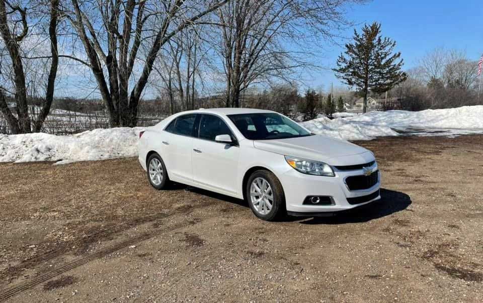 White sedan parked on a dirt lot, snow in the background, trees, sunny day.