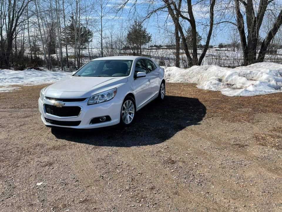 White Chevrolet Malibu parked on a gravel surface, with snow and trees in the background.
