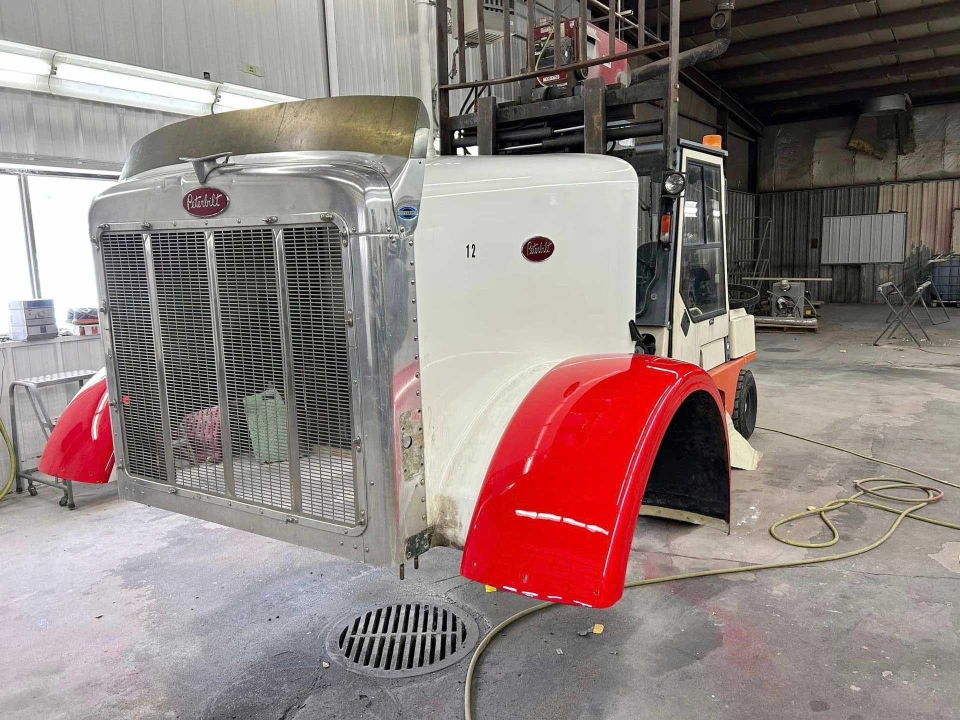 A semi-truck front end on a forklift inside a workshop; chrome grill, red fenders, white hood.