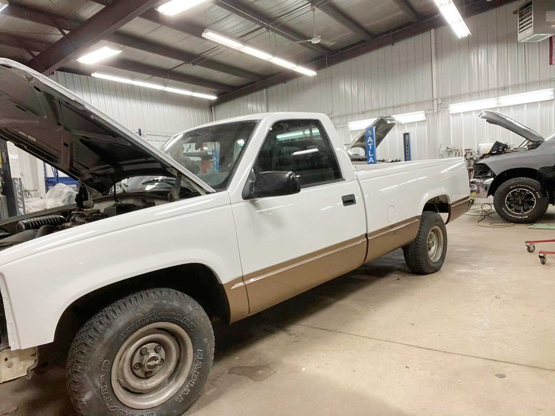 White and brown pickup truck with hood open inside a repair shop.