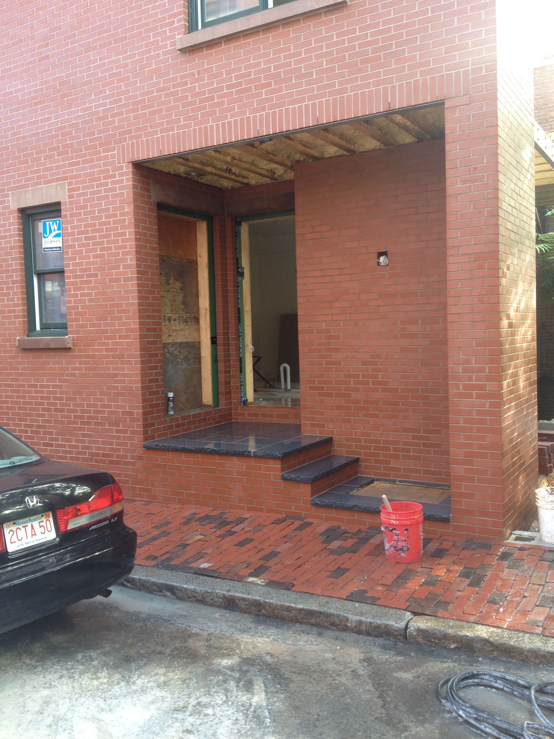 Exterior view of a brick house entryway featuring stone steps, an unfinished doorway, and a red bucket on the ground.