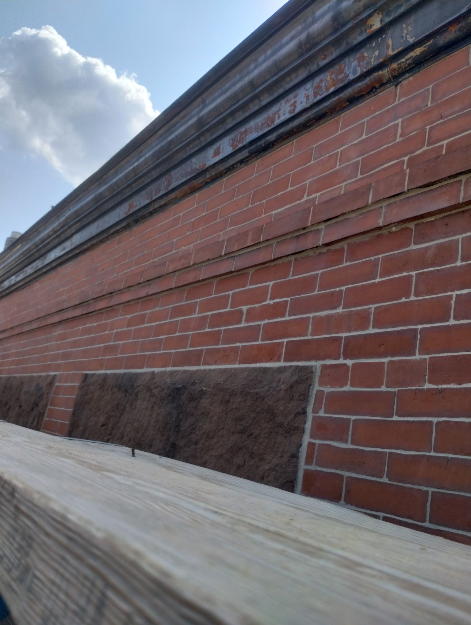 A low-angle view of a red brick wall with dark stone accents below a dark metal ledge under a blue, cloudy sky.
