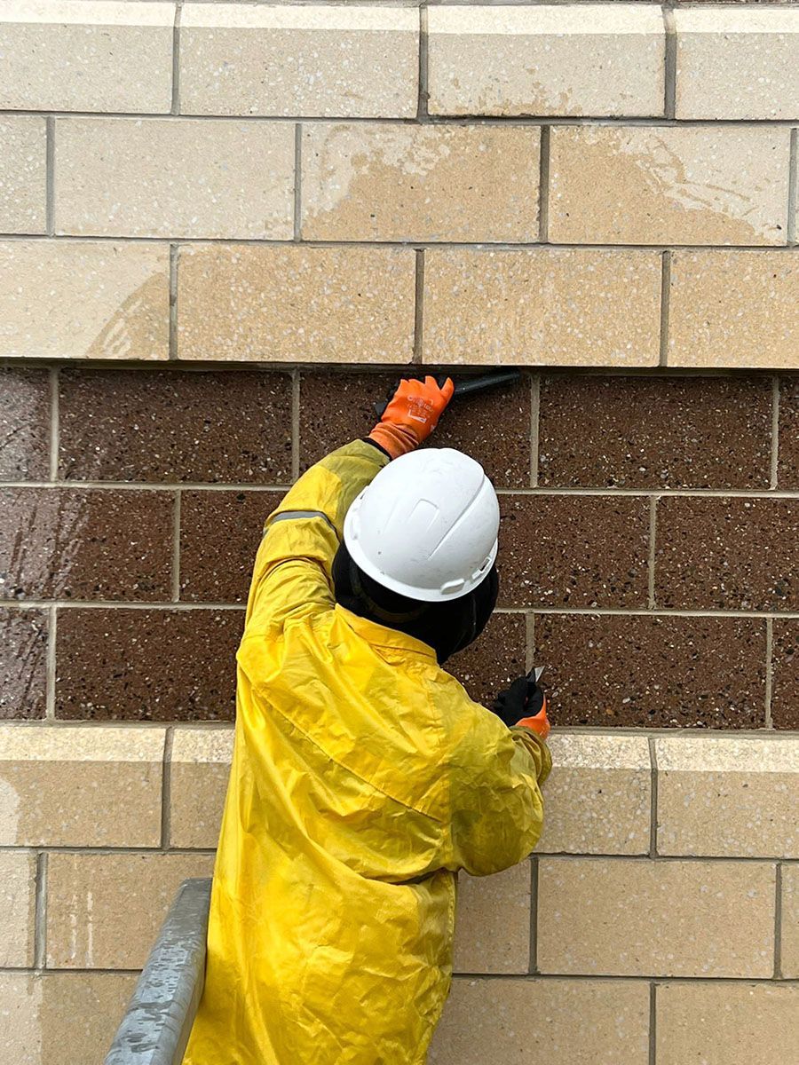 A man in yellow PPE and a white hard hat is cleaning a brick wall