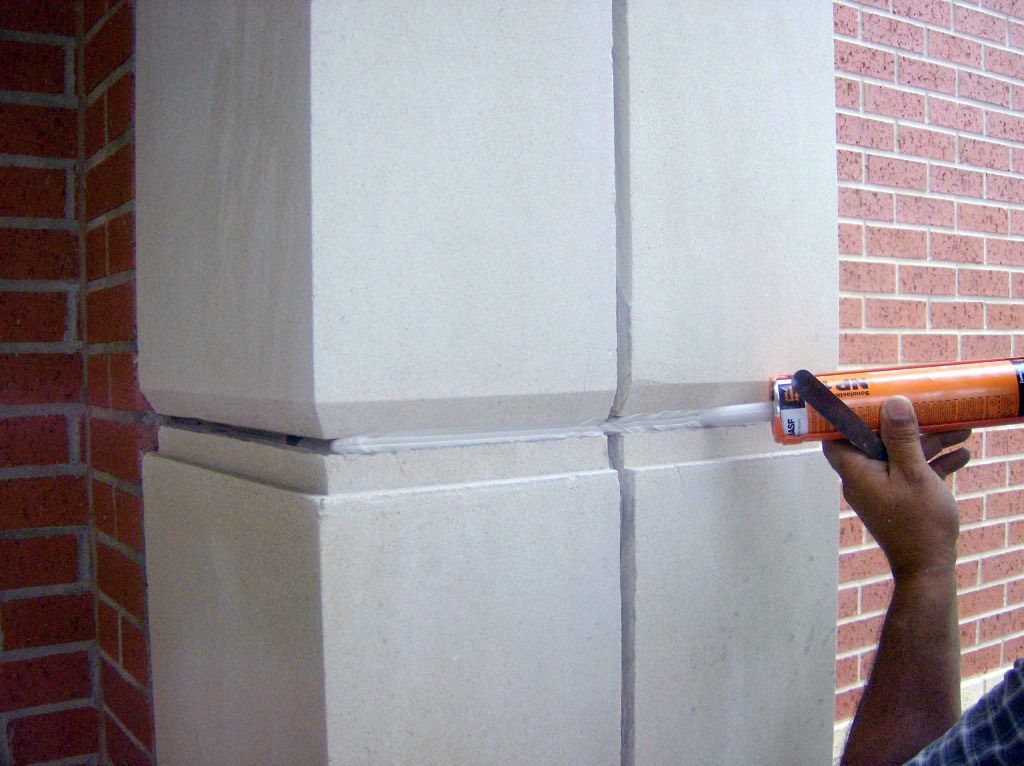 Person caulking a rectangular stone pillar joint with a caulk gun near a brick wall.