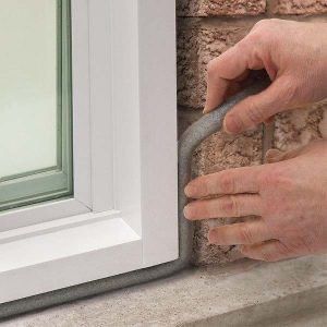 Hands applying gray foam weather stripping around a window frame and brick exterior.