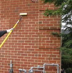Person painting a brick wall with a paint roller on a long handle, outdoors.