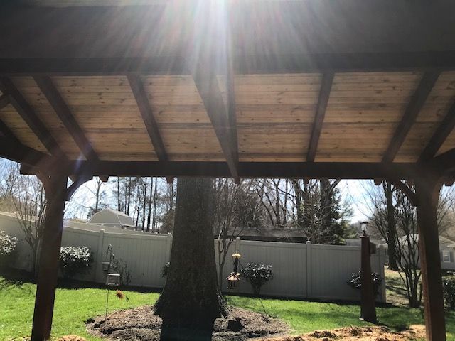 Wooden pavilion with a large central tree, grassy yard, and fence in bright sunlight