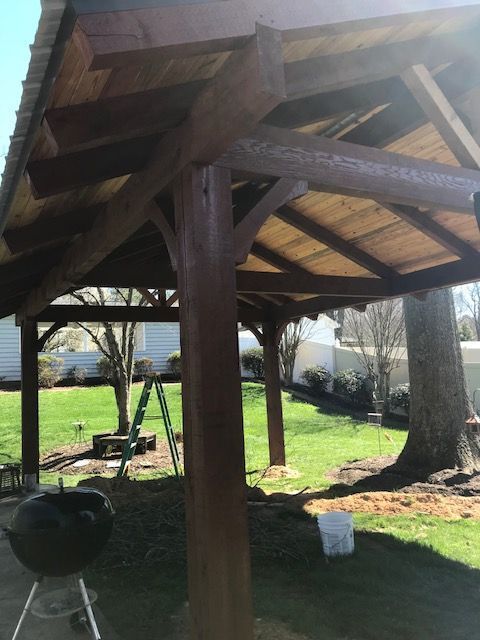 Wooden pavilion roof and support posts in a grassy yard, with a charcoal grill nearby.