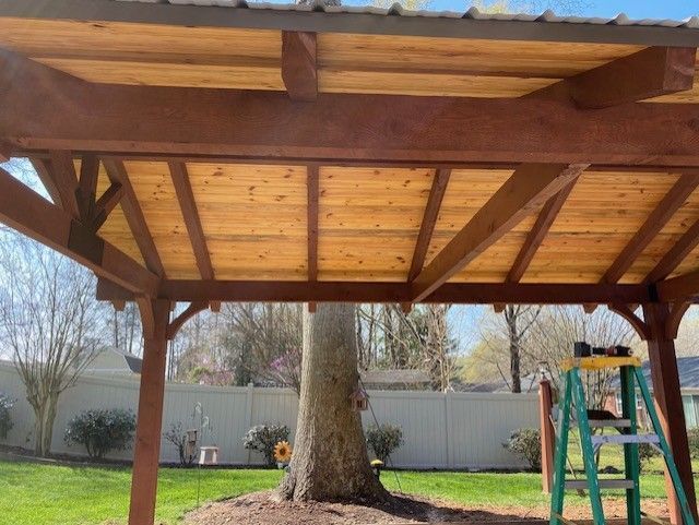 Wooden backyard pavilion with a large tree trunk centered beneath it and a white fence in the background