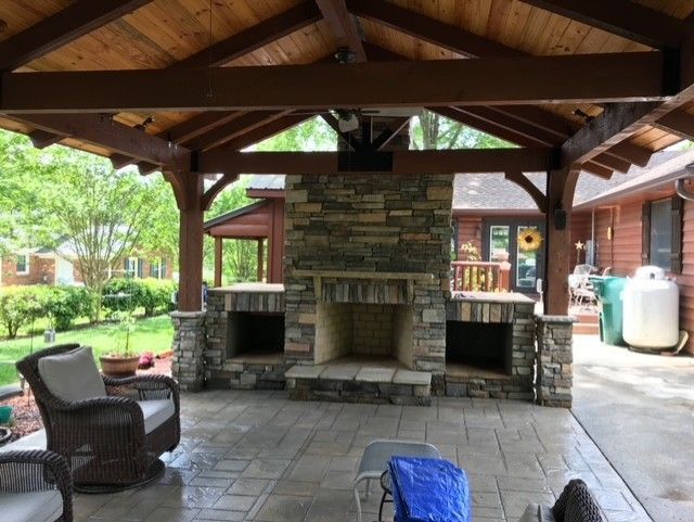 Covered stone outdoor fireplace beneath a wooden pavilion with patio chairs and garden surroundings