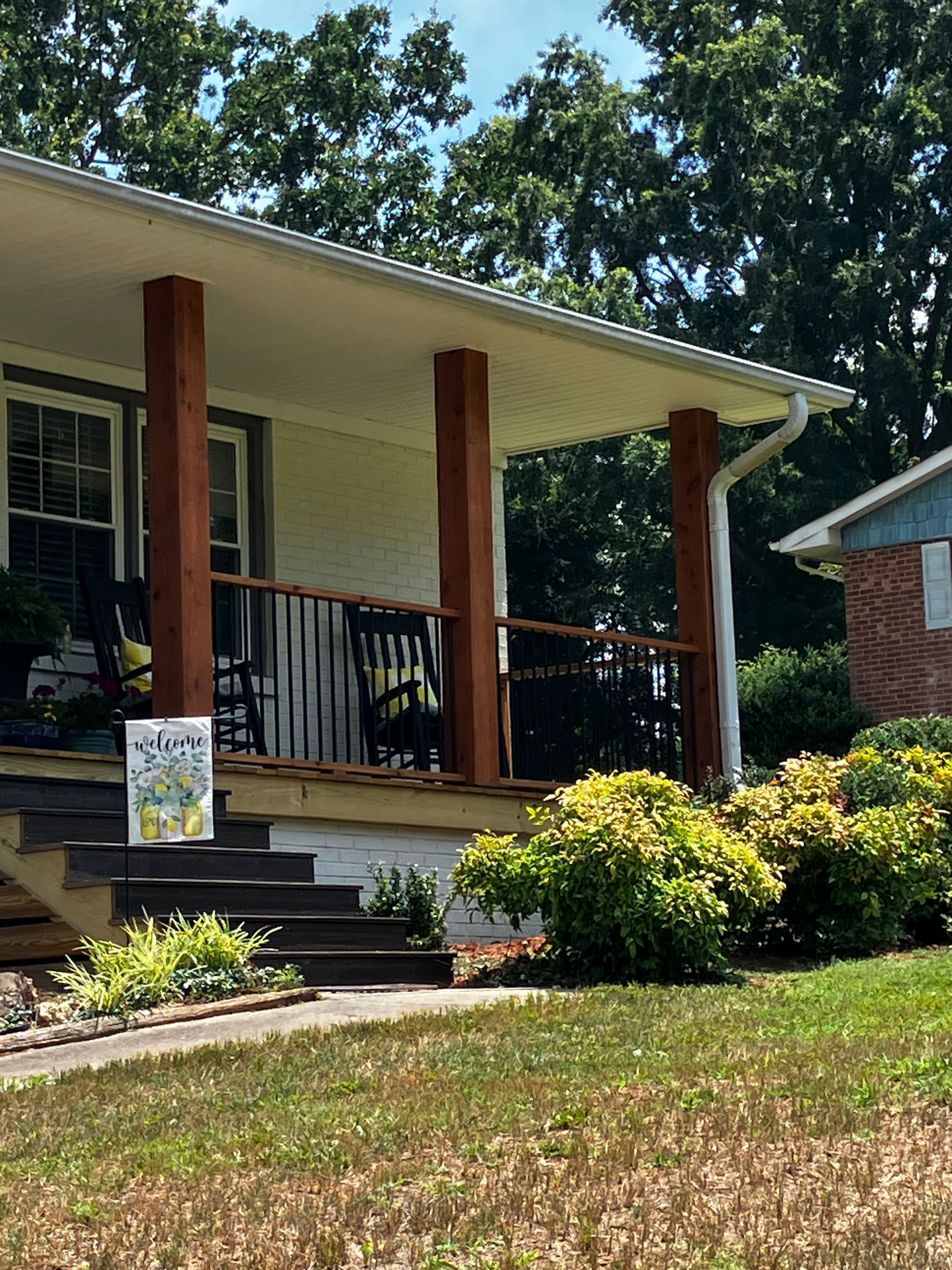 Front porch of a house with white siding, brick columns, black railing, and a small sign by the steps