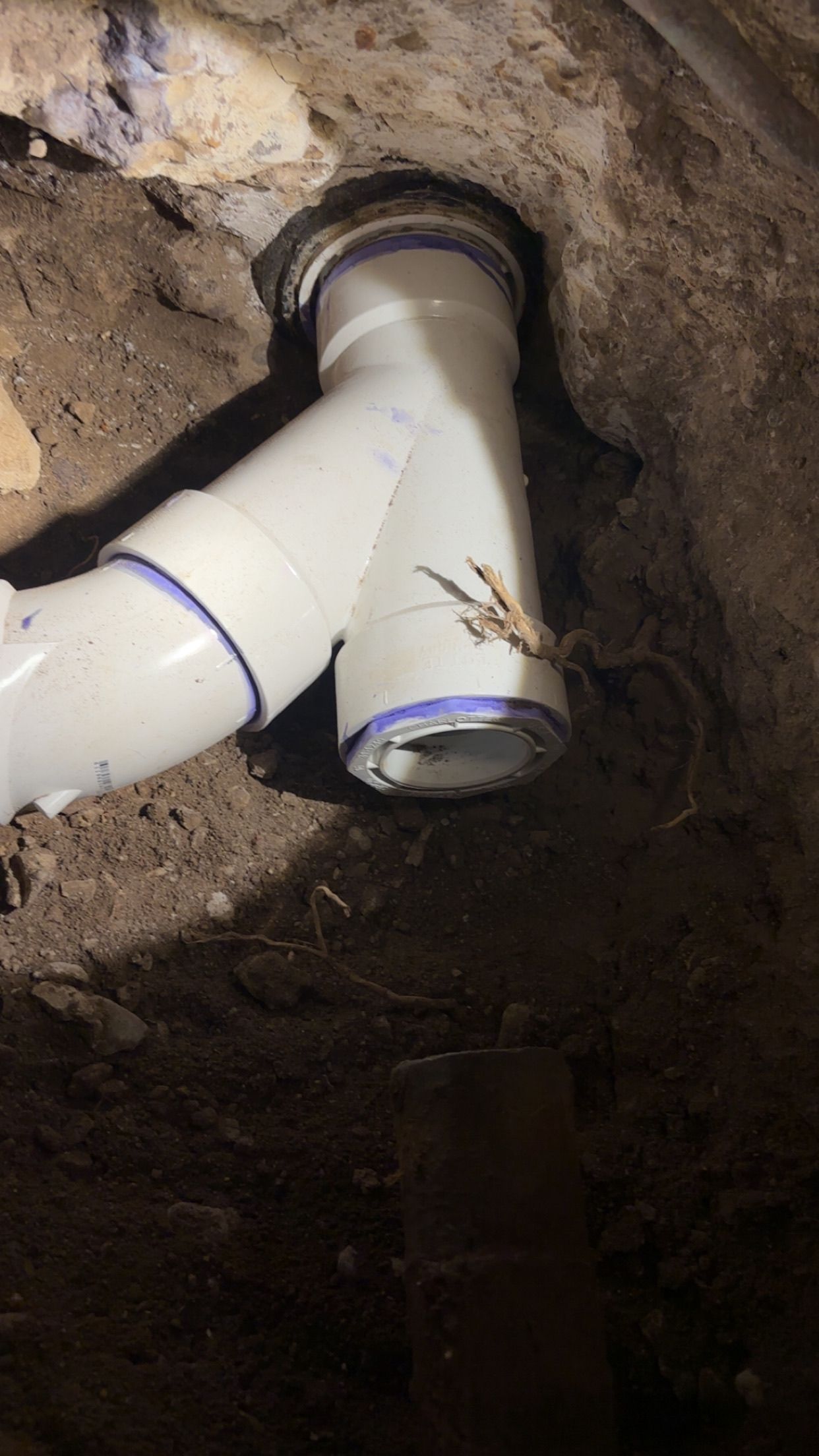 White PVC plumbing pipes and fittings in a dirt trench, near a foundation.