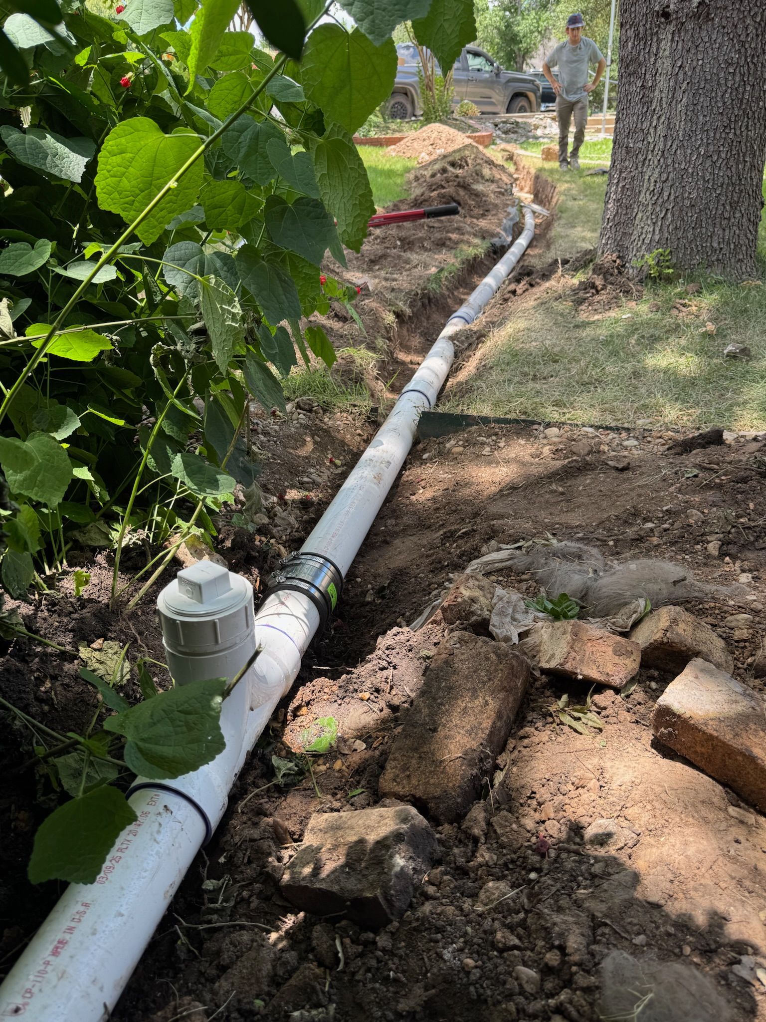 A trench with white pipes is being installed in a yard; a person stands nearby.