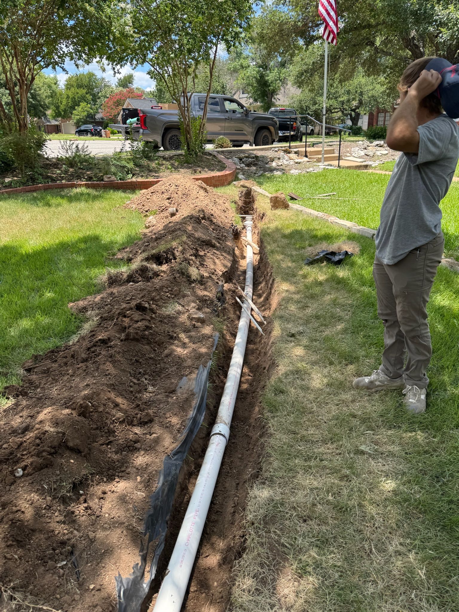 Man carrying a water tank next to a trench with a white pipe running through it in a lawn.