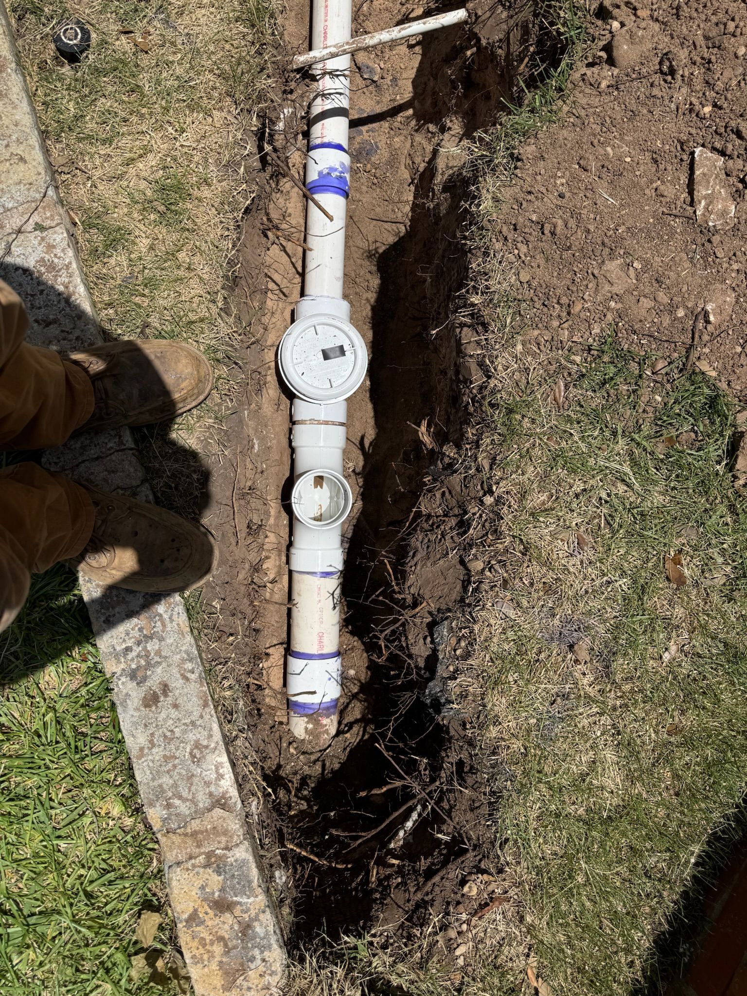 Irrigation system pipe in trench next to lawn and brick border. White pipe with pressure gauge and blue fittings.