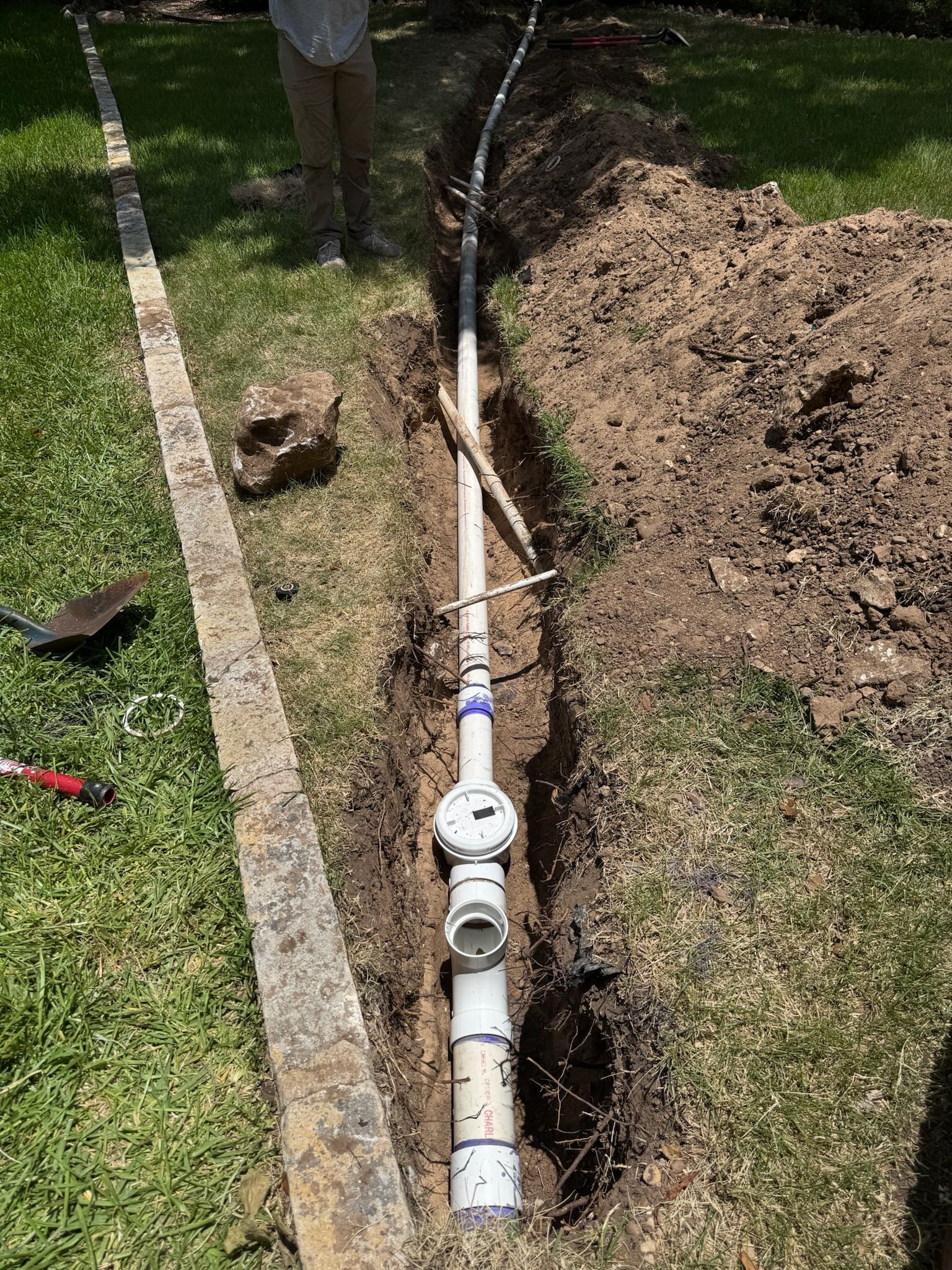 A trench with a white pipe and water meter, in grass next to a paved area; a person stands nearby.