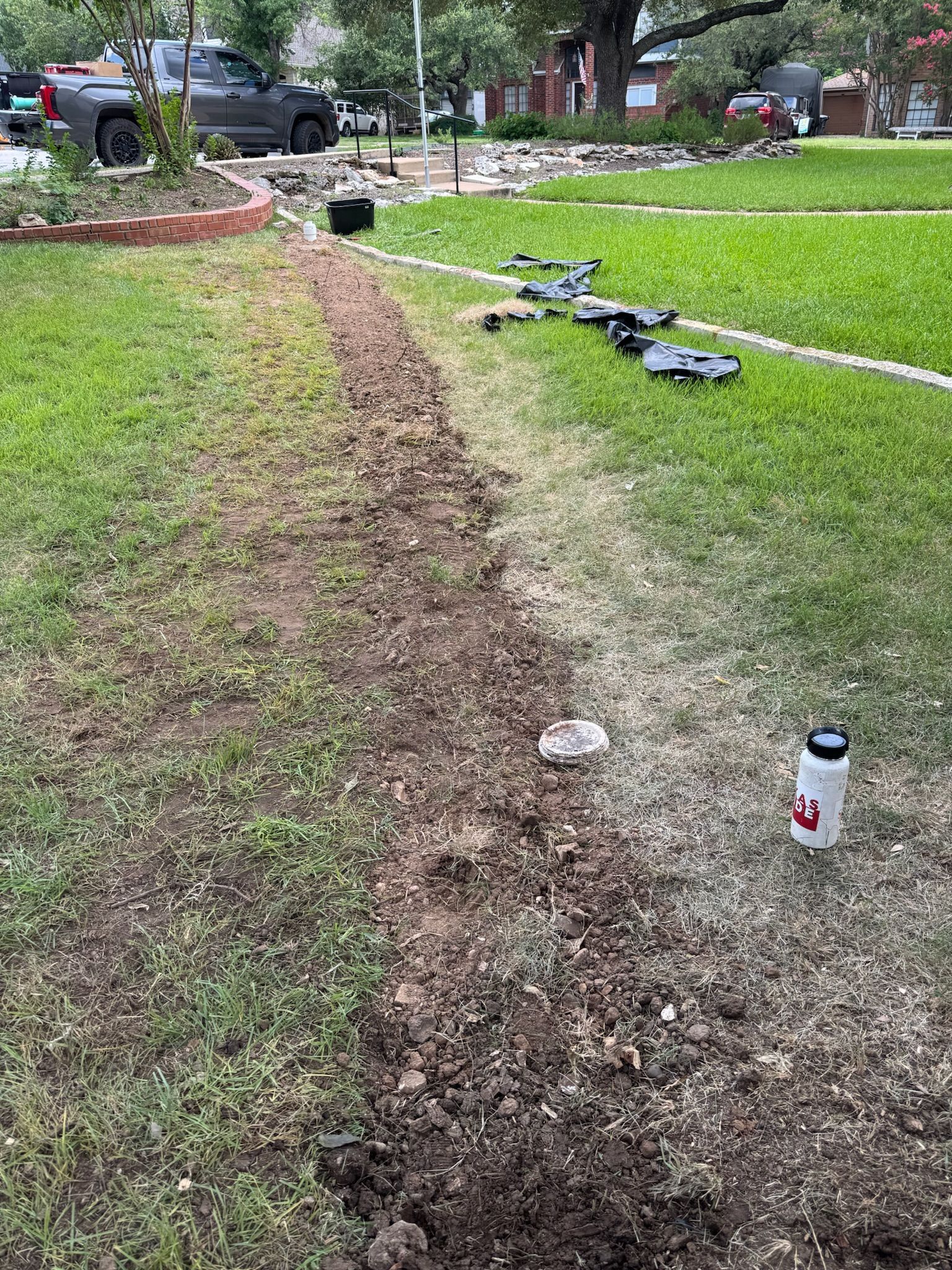 Trench dug in grass for irrigation system installation, tools and water bottle lie nearby.