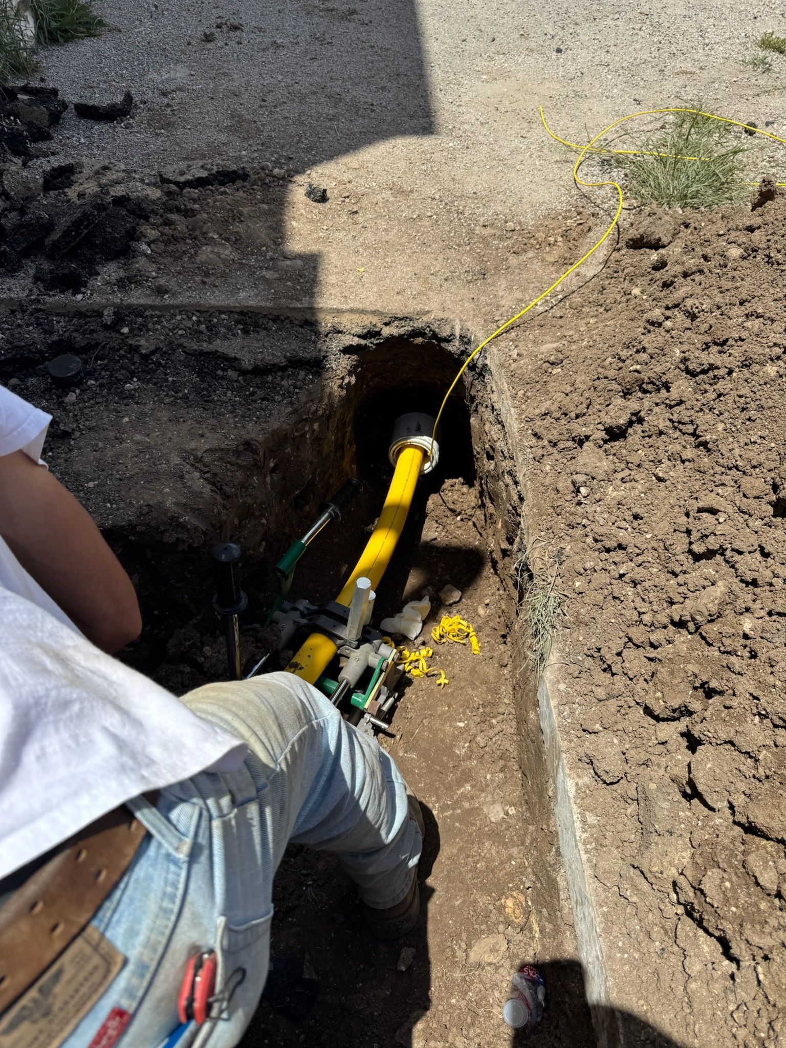 Person in jeans working on yellow gas line in a trench. Yellow cable and dirt visible.
