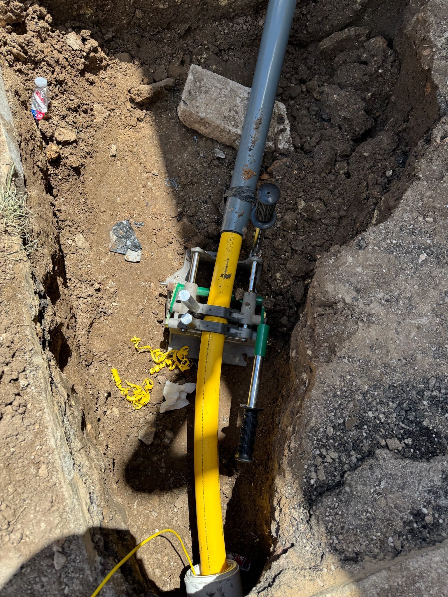 Yellow gas pipe being worked on in a trench. The pipe is clamped with tools and surrounded by dirt and asphalt.