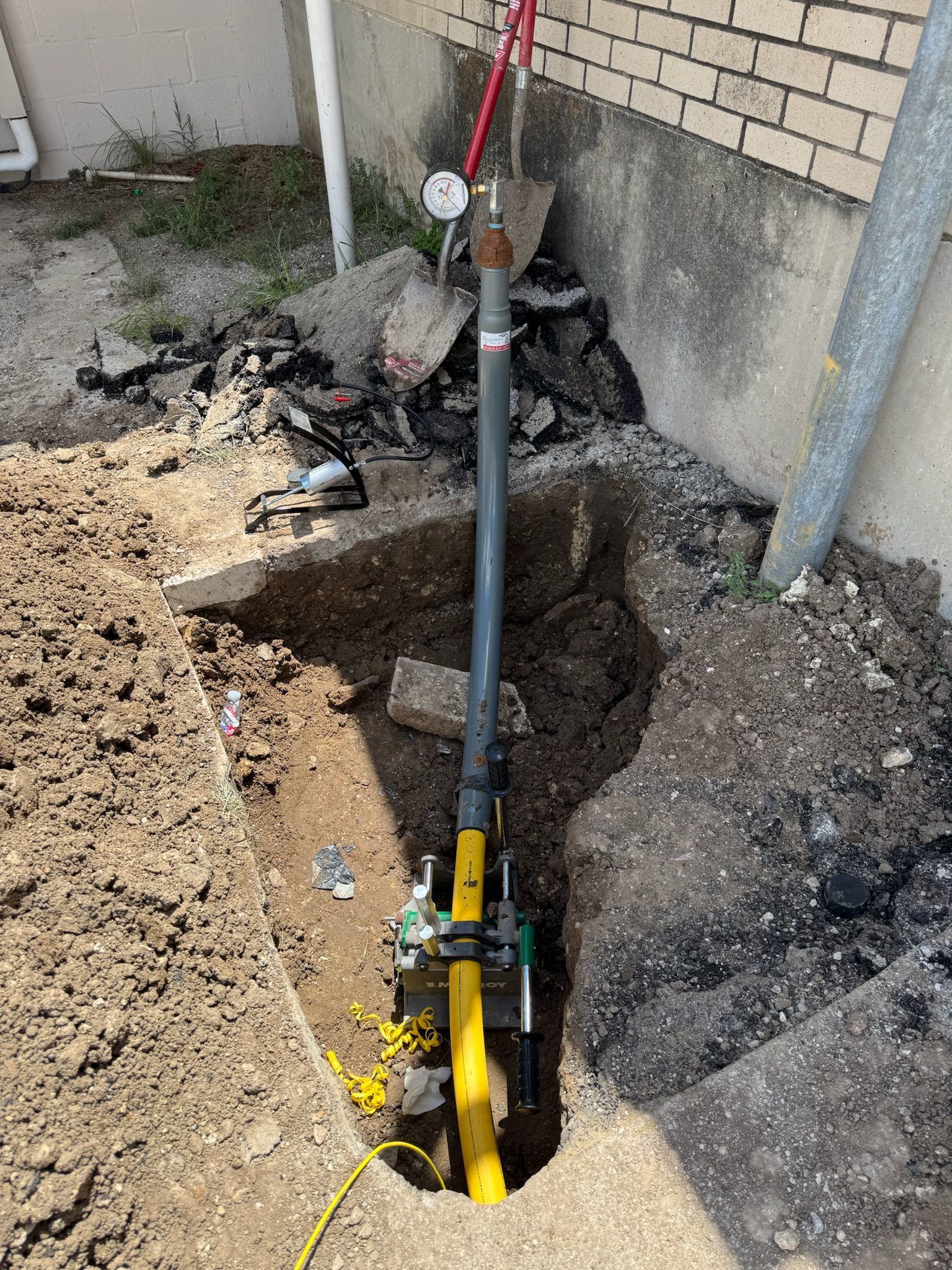 A gas line repair in progress; a trench dug around yellow pipes, grey pipe rising, pressure gauge attached.