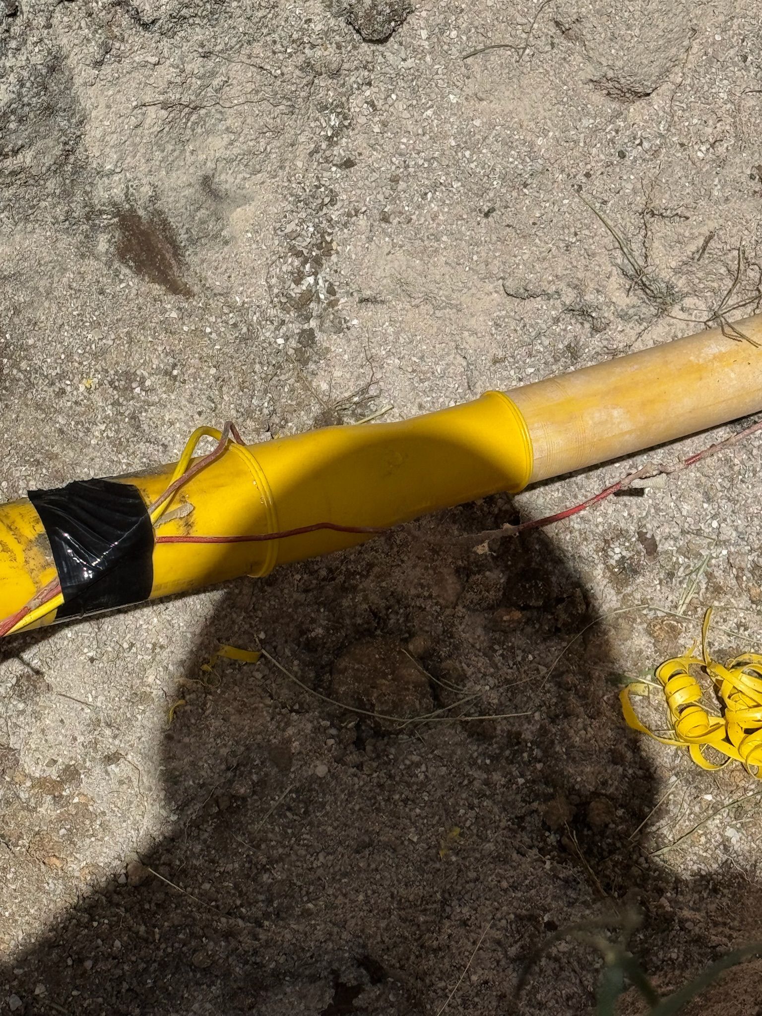 Yellow pole with black tape and red wire on concrete with dark shadow and brown debris.